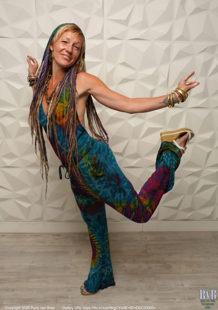 Woman with long hair in colorful outfit poses with one leg raised and arms in peace signs, smiling, against a textured wall.