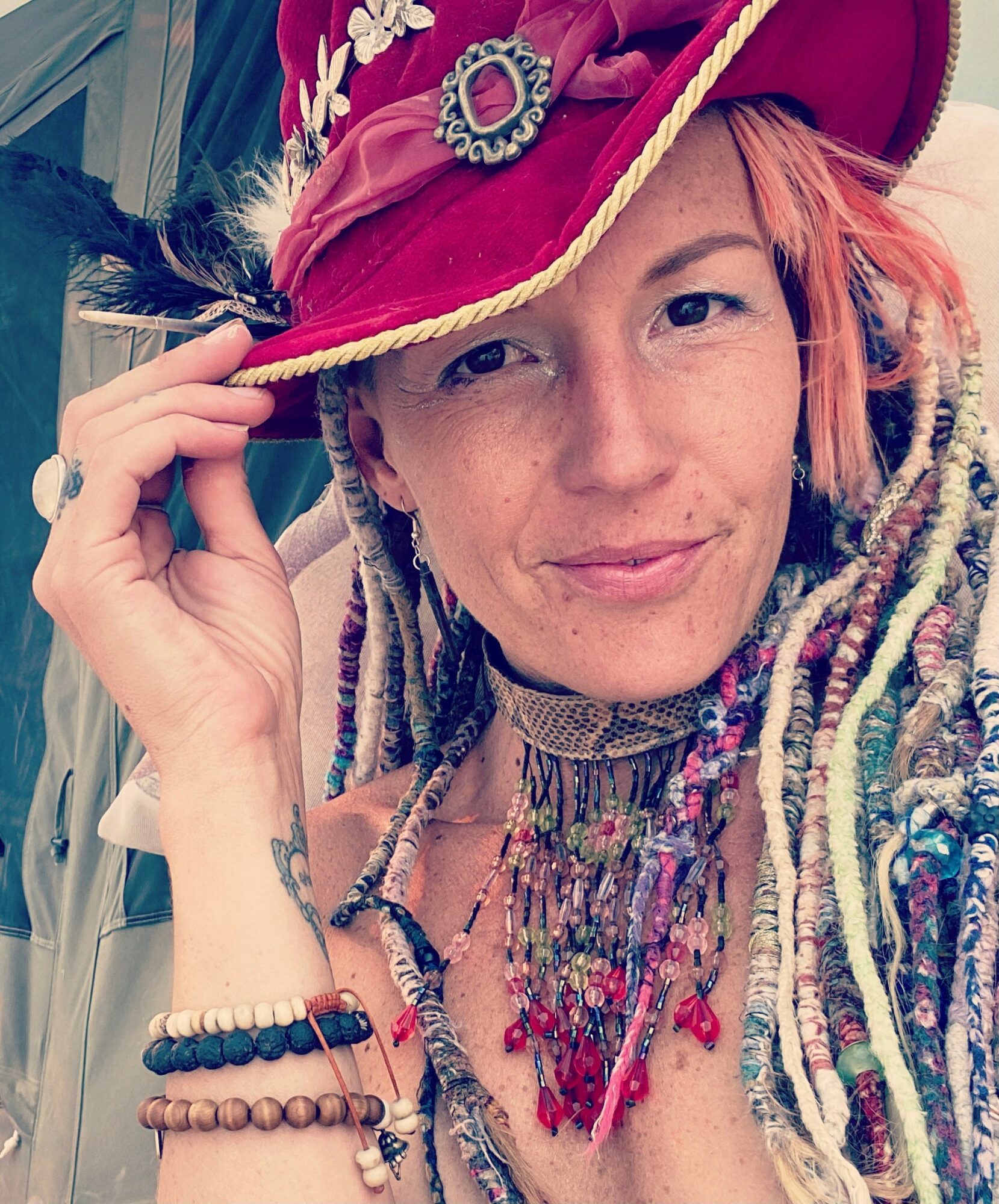 Woman wearing a colorful hat with feathers, beads, and jewelry, smiling and holding the hat's brim.