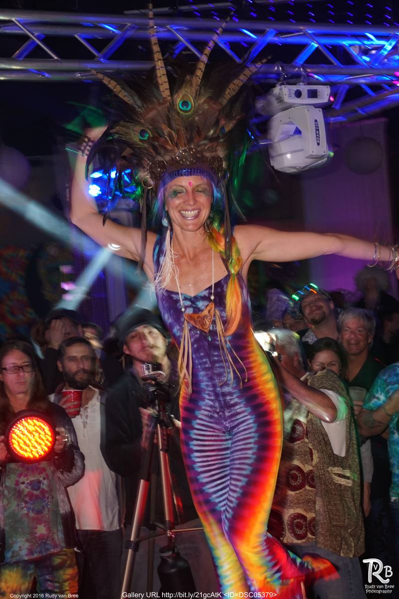 Performer in colorful costume on stage with audience, smiling, with bright lights and equipment overhead.