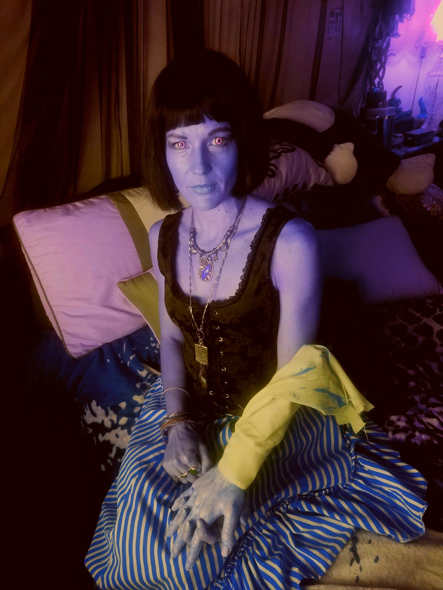 Woman with dark hair and makeup sitting on a bed, wearing a black dress and jewelry, with a yellow jacket and striped skirt.