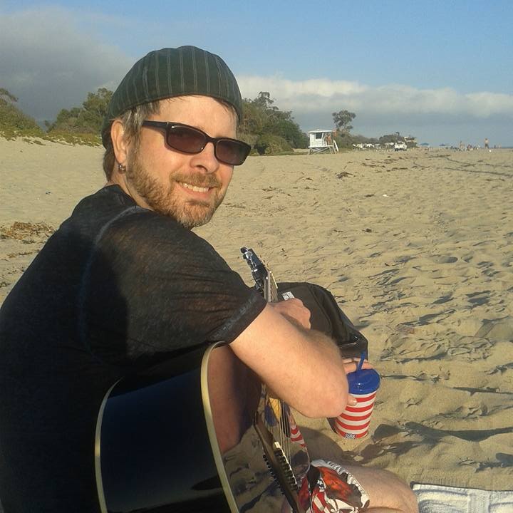 Man sitting on sandy beach holding a guitar, wearing sunglasses, a beanie, and a black shirt, with a blue sky and distant trees.