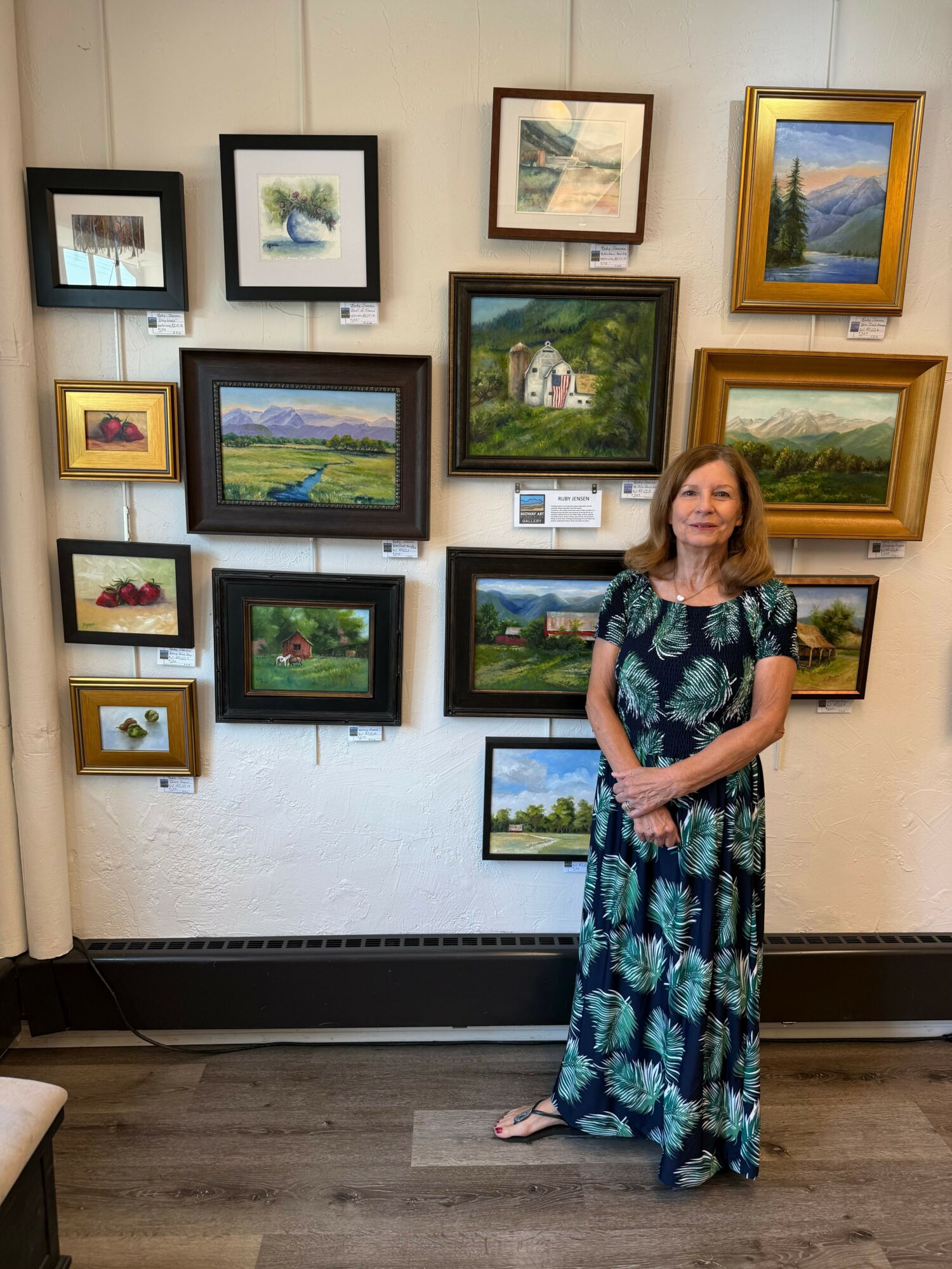 Woman in a patterned dress standing in art gallery with framed landscape paintings on wall.