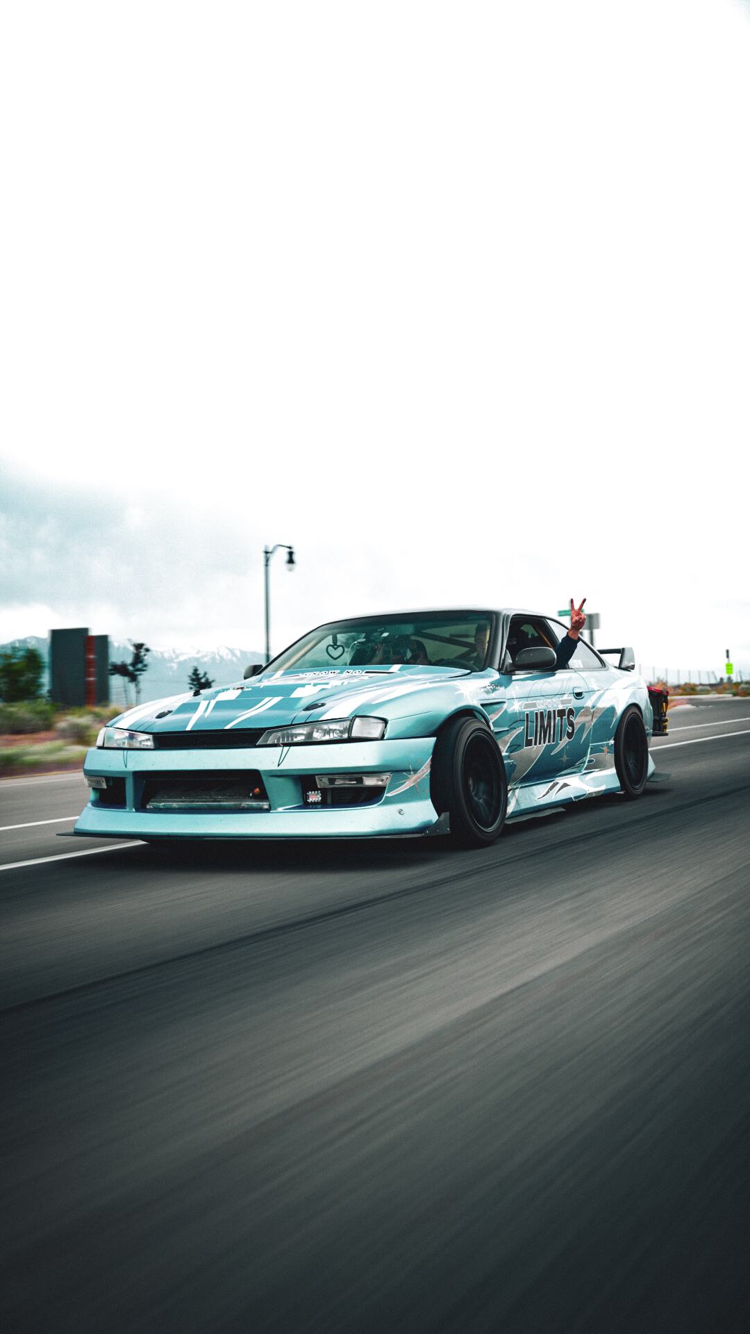 A light blue sports car driving on a road with a cloudy sky in the background.