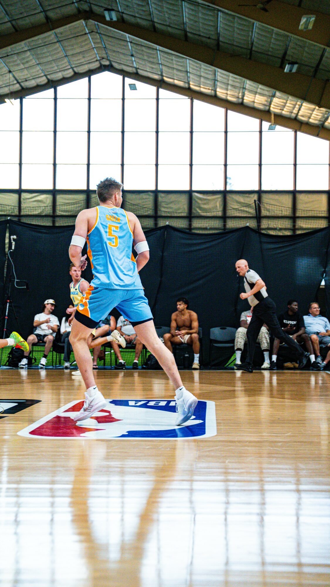 Basketball player in blue jersey jumps on court with spectators and referee in background.