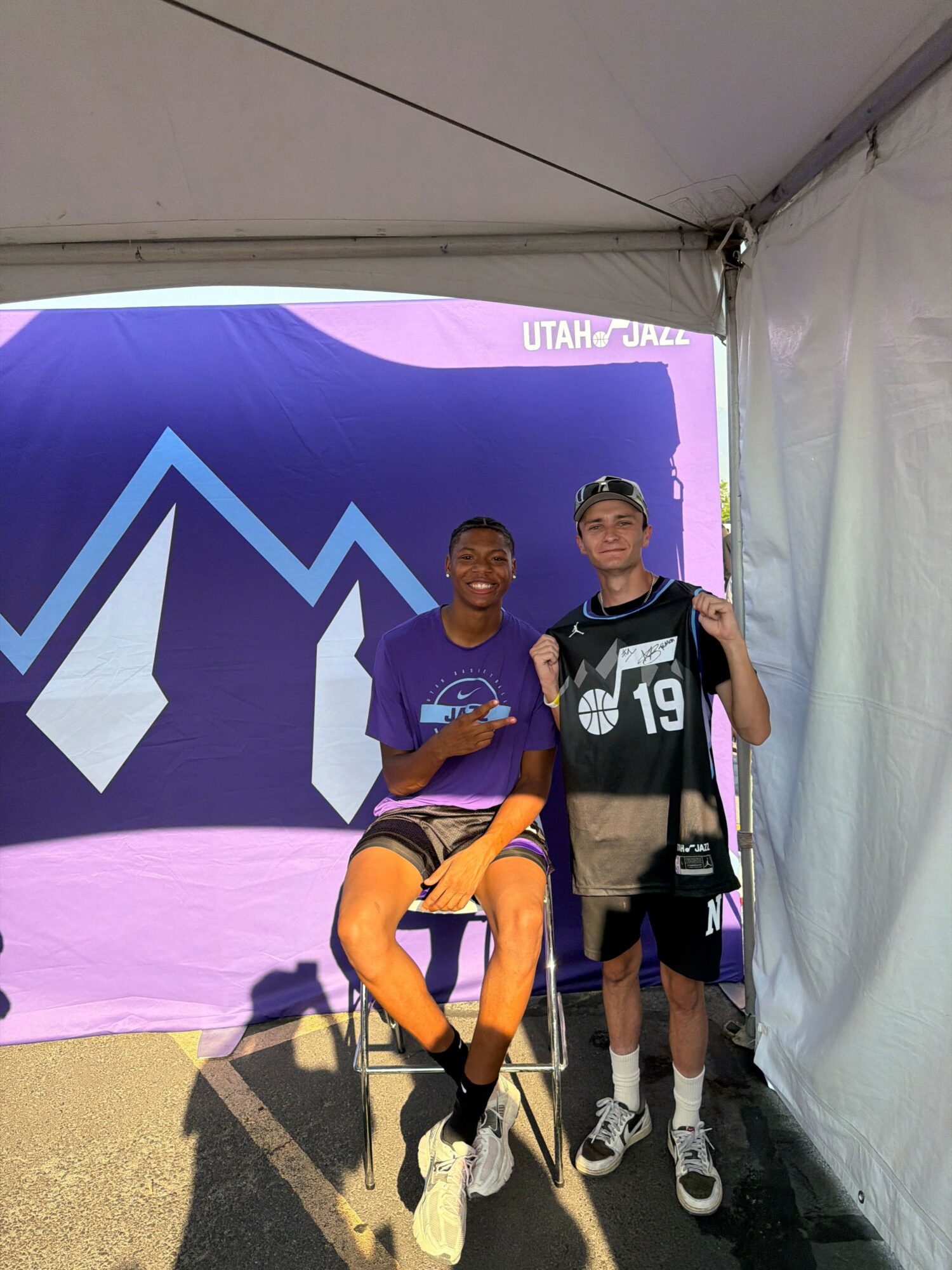 Two athletes stand in front of a purple and blue backdrop with a large white arrow and the text 'ULTRA TRAIL'. One is standing, the other is sitting on the ground, both wearing sports gear and bib numbers.
