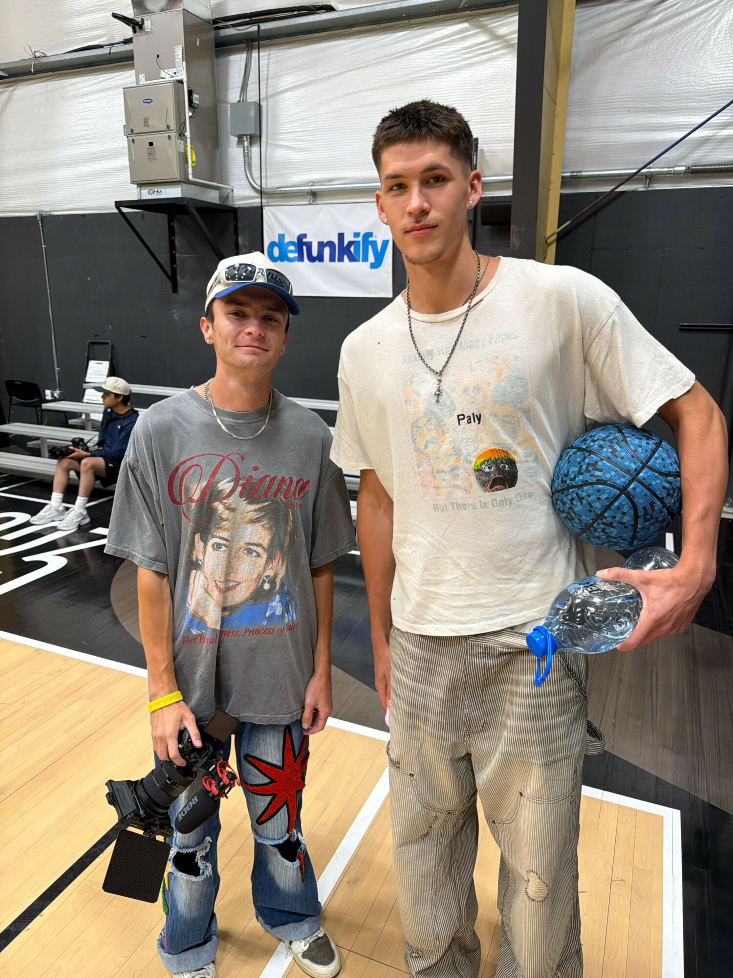 Two young men standing in a gym, one holding a water bottle, the other with a camera, both smiling.