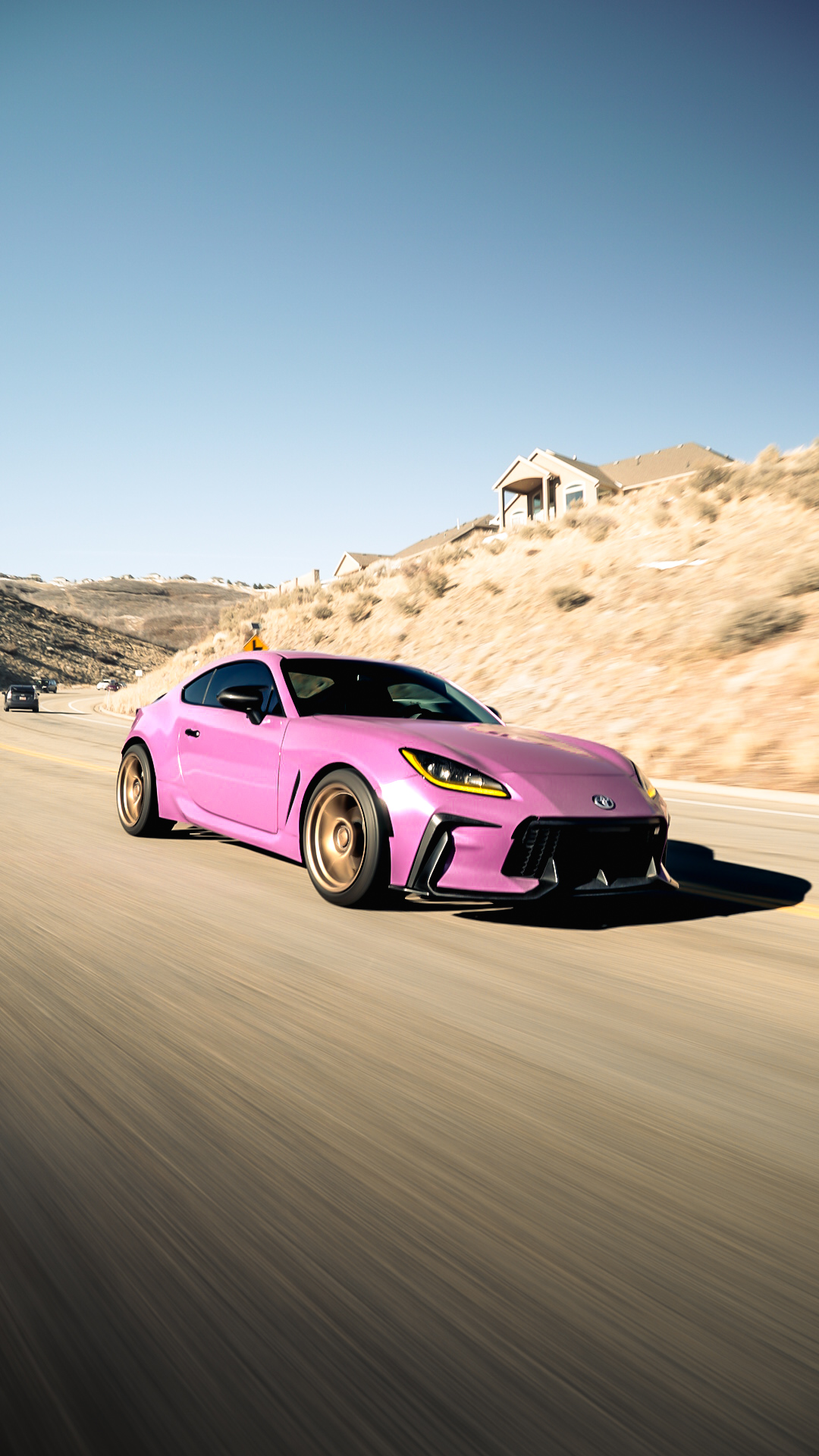 Pink sports car driving on a road with a dry, hilly landscape and a house in the background under a clear blue sky.