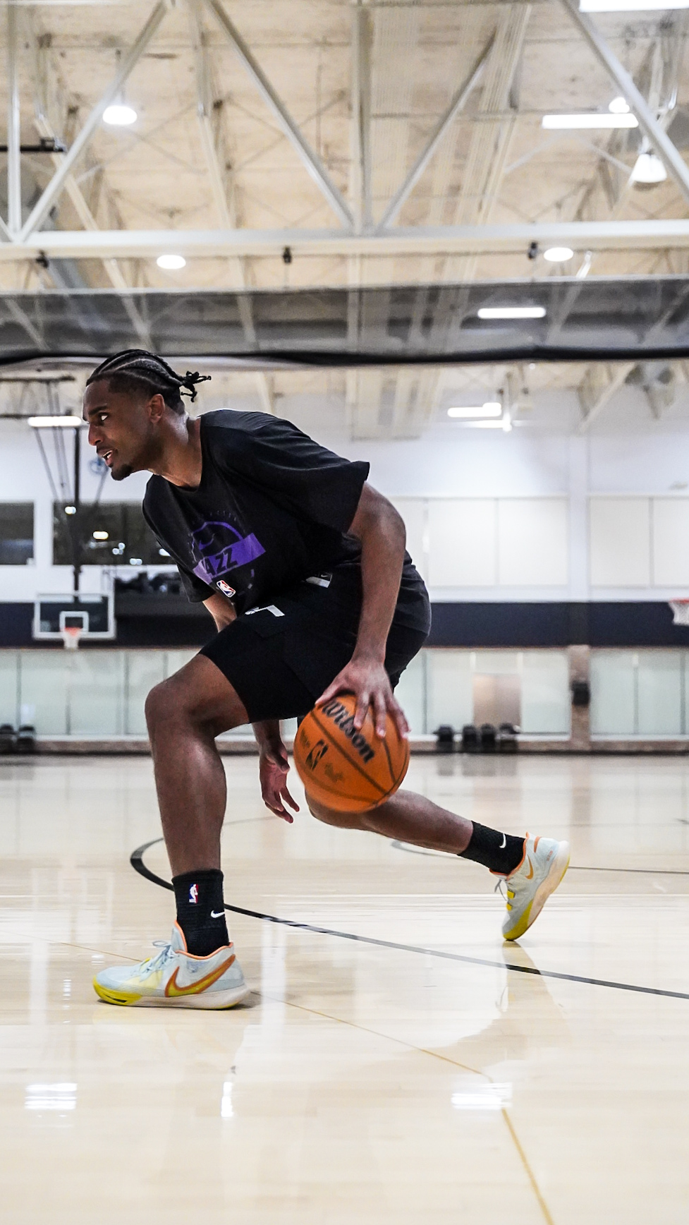 Person dribbling basketball on indoor court, wearing black shirt, shorts, and sneakers, with gym ceiling overhead.