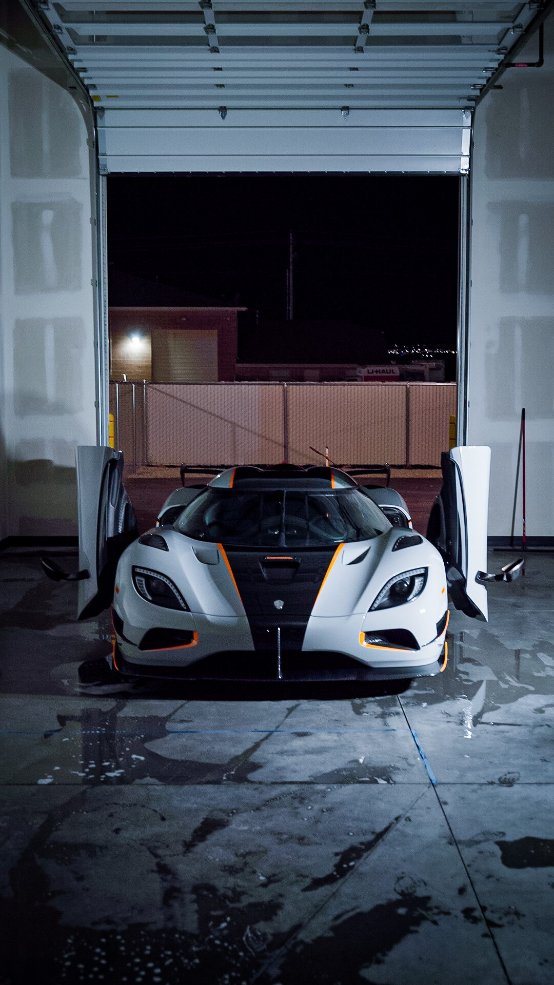 Sports car parked inside a garage with open door, wet floor, and city lights visible outside.