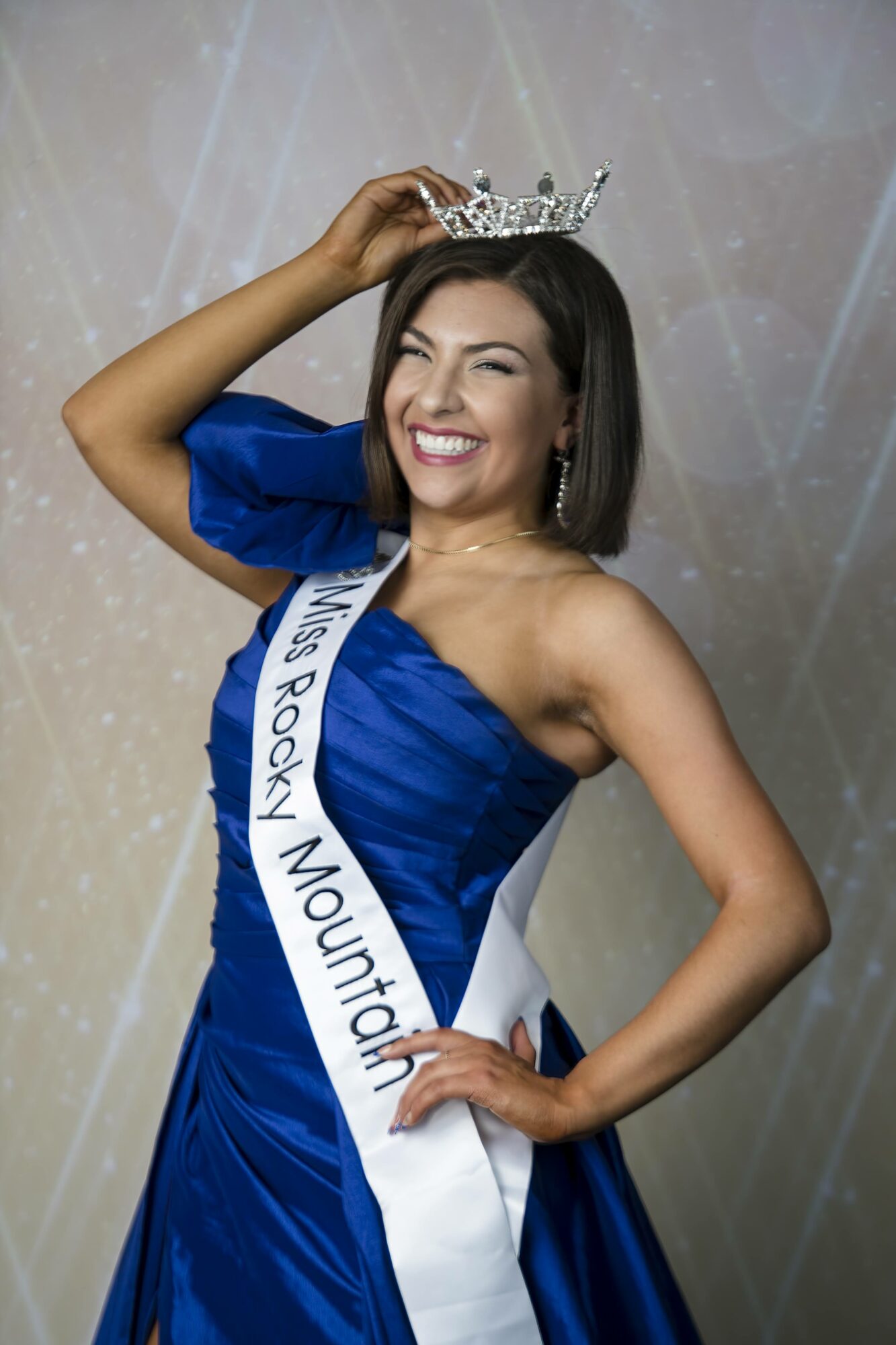 Woman wearing a blue dress, crown, and sash, smiling, with one hand on her hip and the other adjusting her crown.