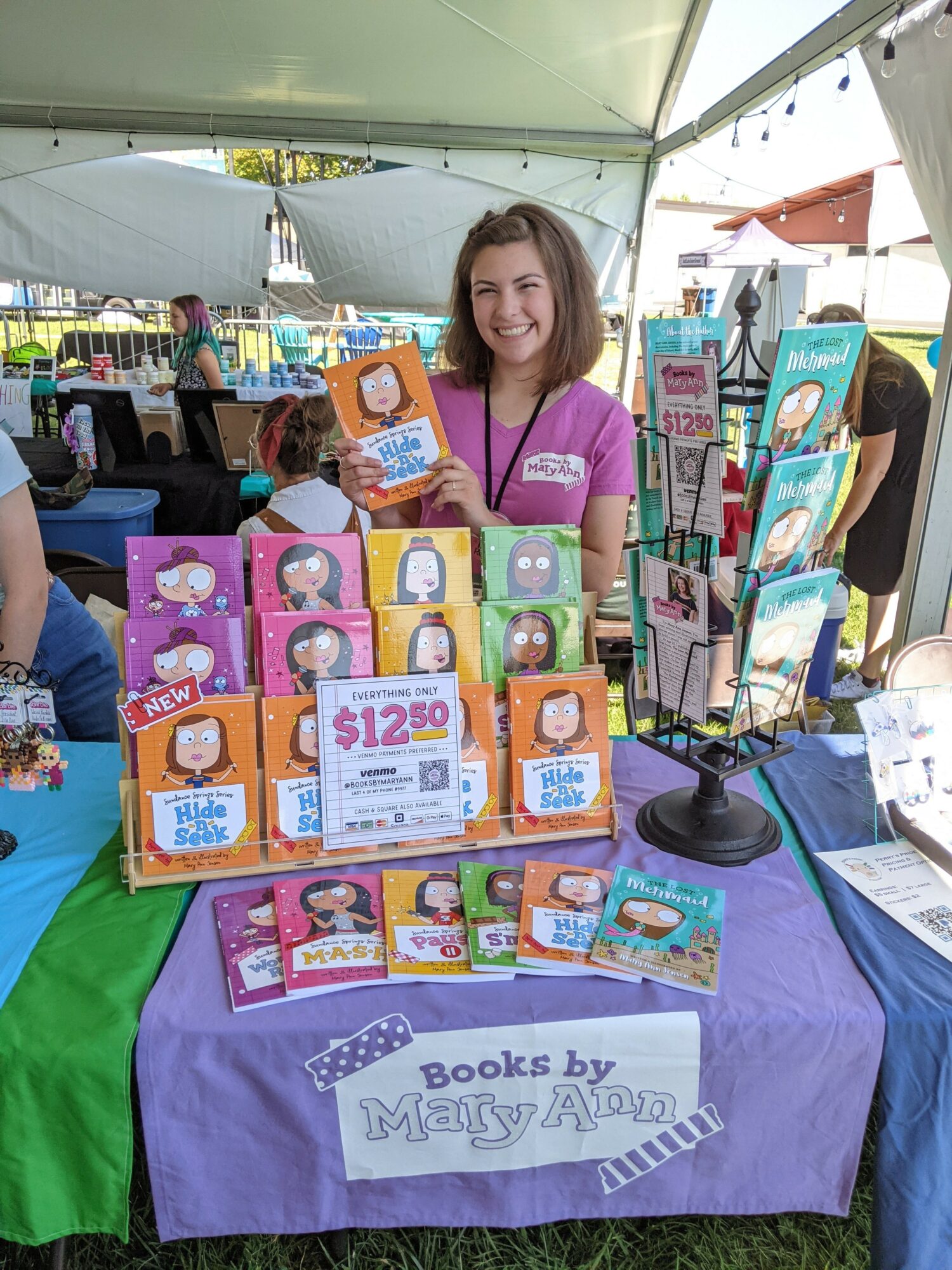 Smiling woman at book booth with colorful books and display stand outdoors under a canopy.