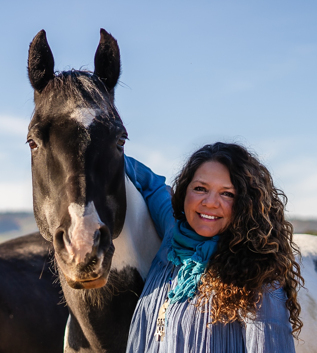 Woman with curly hair smiling next to a black and white horse outdoors against a blue sky.
