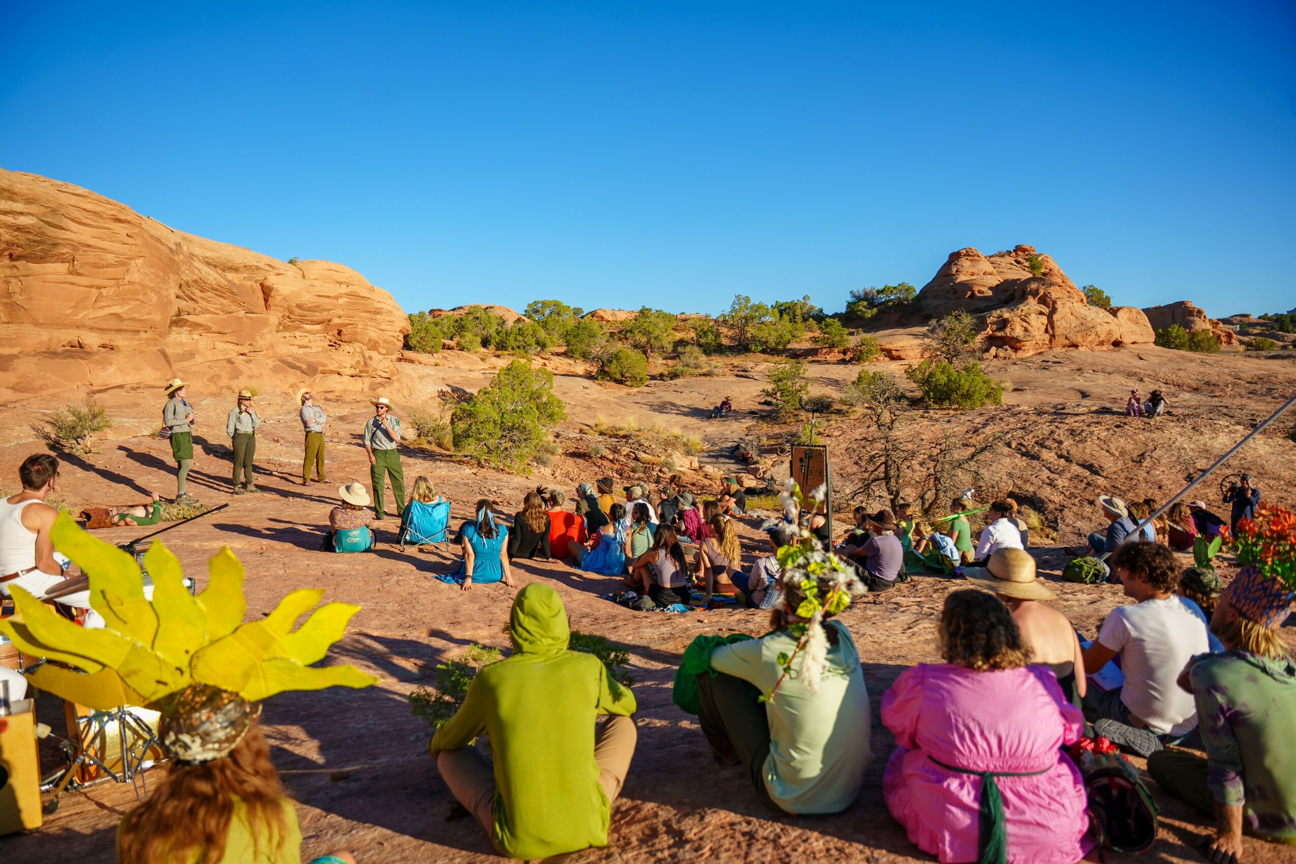 Group of people sitting outdoors on rocky terrain with red rock formations and a clear blue sky.