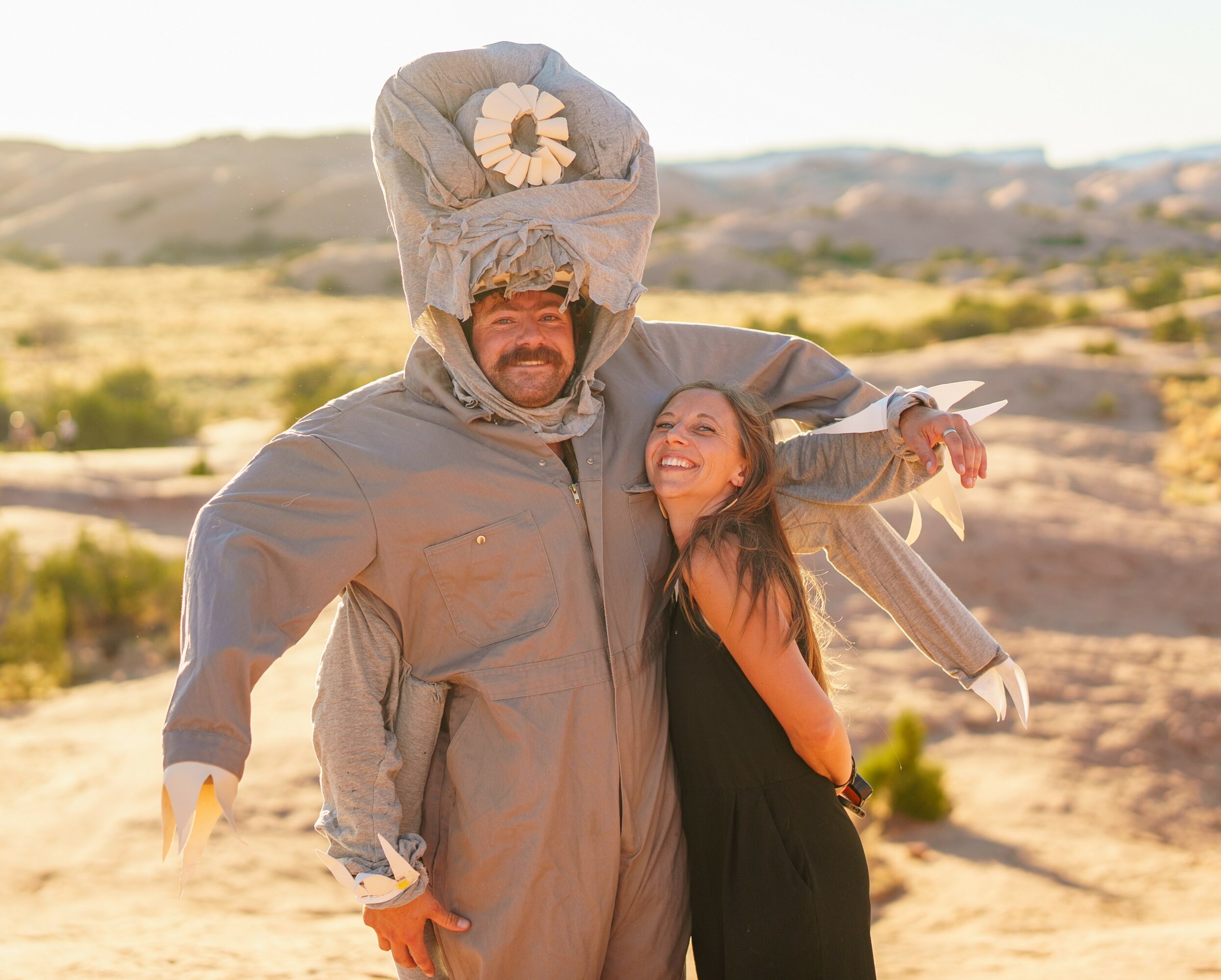 Man and girl in desert landscape, man wearing costume with large headpiece, girl smiling, both standing outdoors.