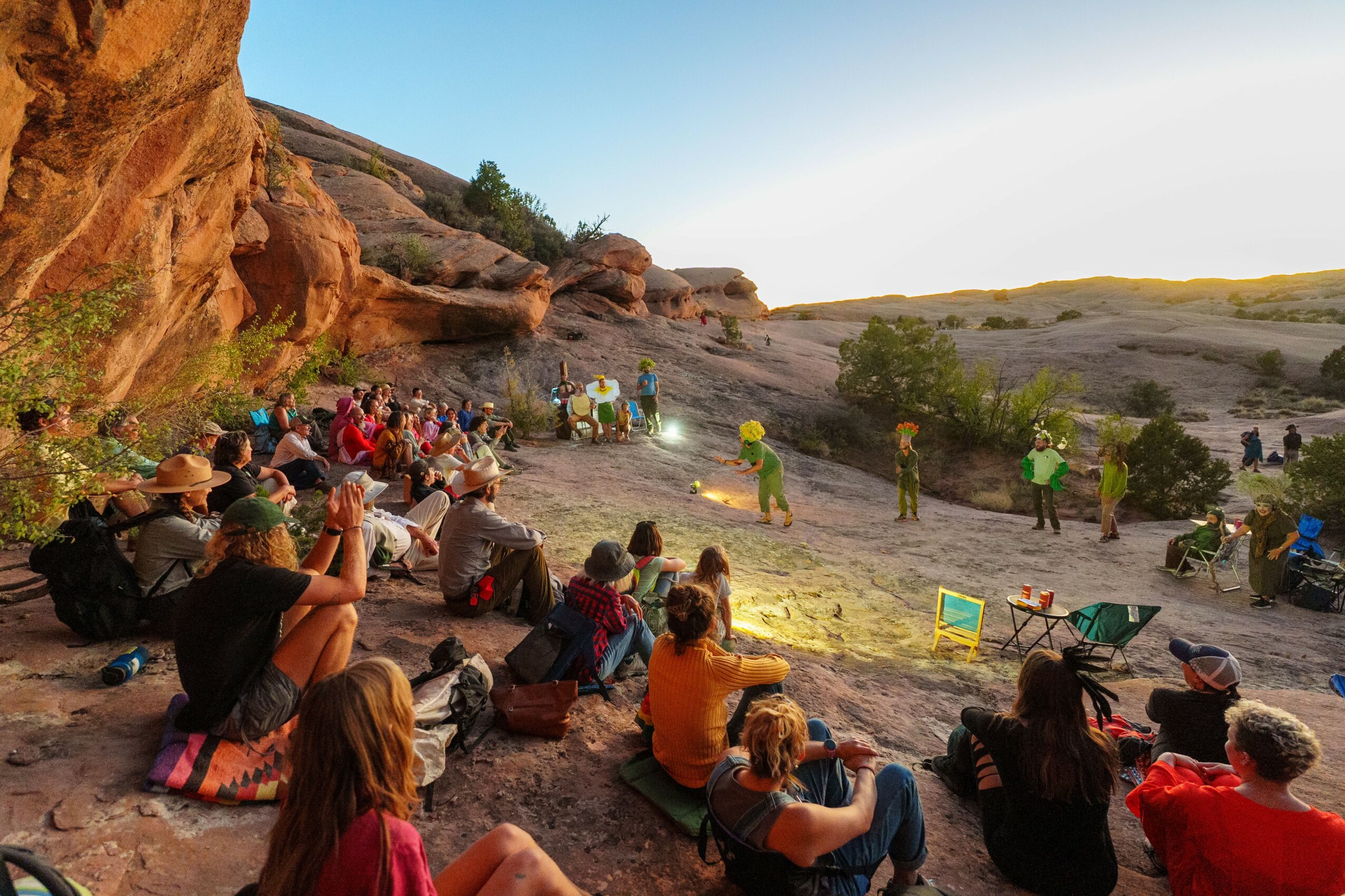 Group of people sitting outdoors on rocks, watching a performance or presentation in a natural landscape with hills and trees.
