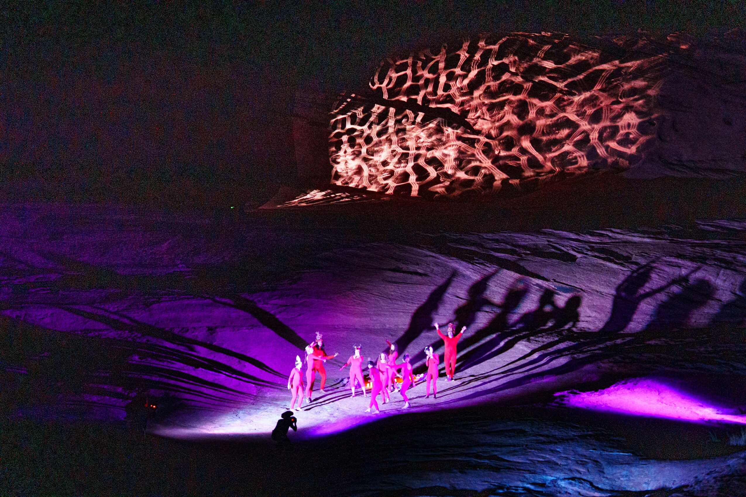Group of performers on stage with colorful lighting and shadow patterns projected on backdrop.