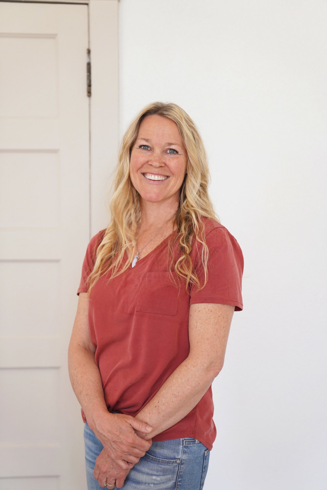 Woman with long blonde hair smiling, wearing a red shirt and blue jeans, standing indoors near a white door.