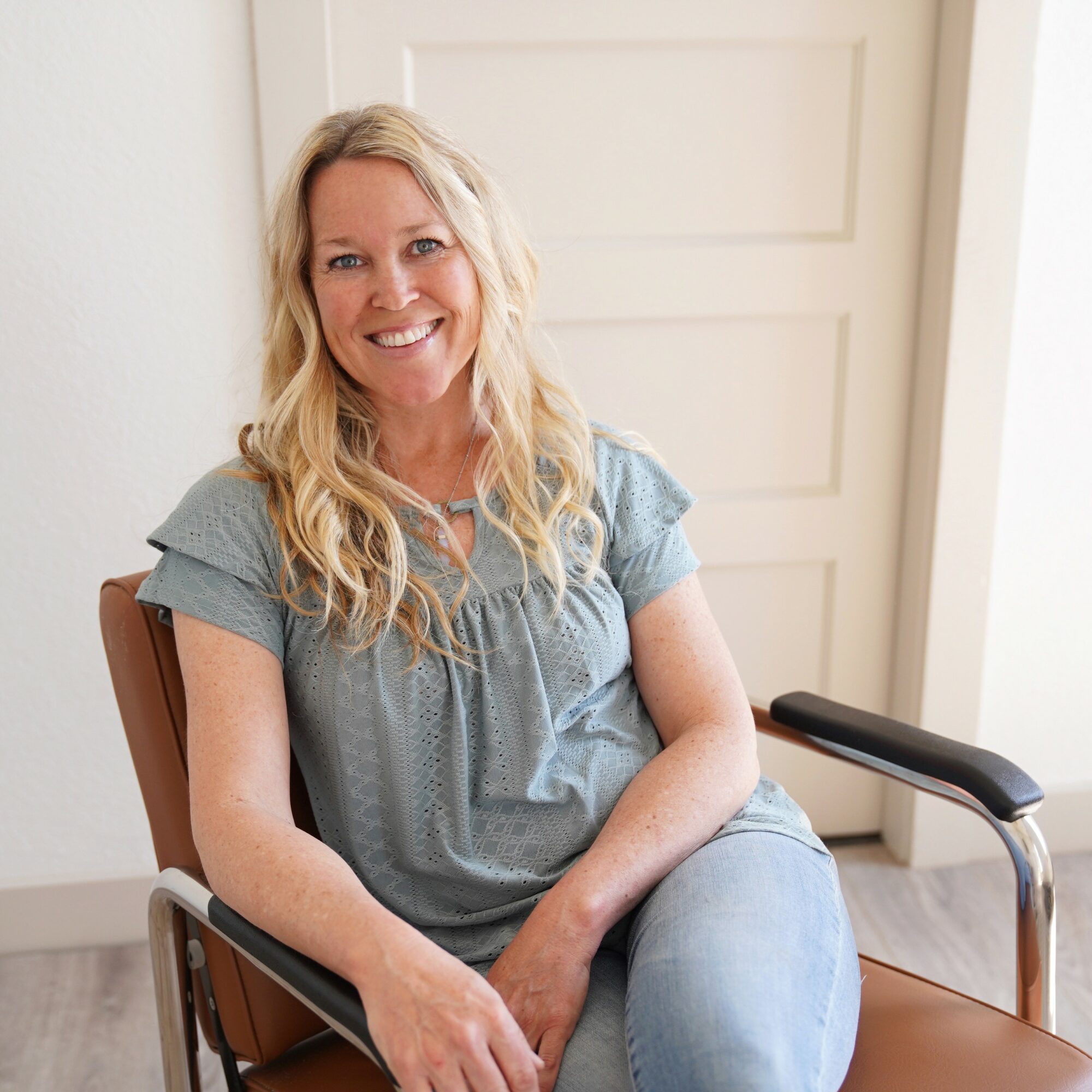Woman with long blonde hair smiling, sitting on a chair indoors, wearing a light blue top and jeans.