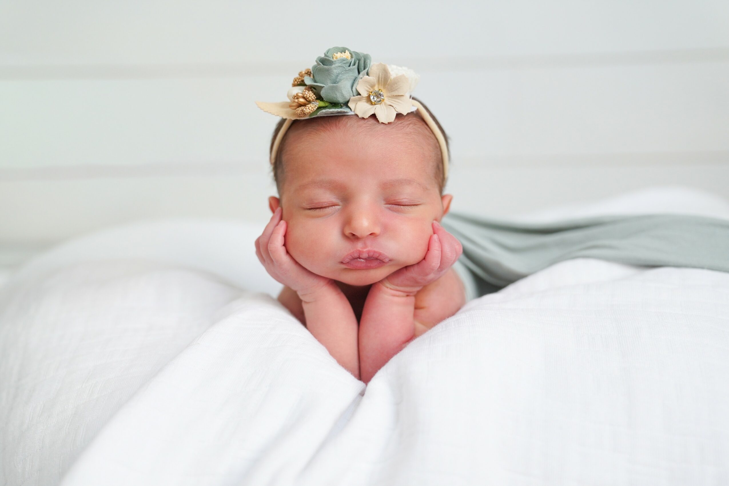 Baby with a floral headband resting head on hands, lying on a white bed, eyes closed, peaceful expression.