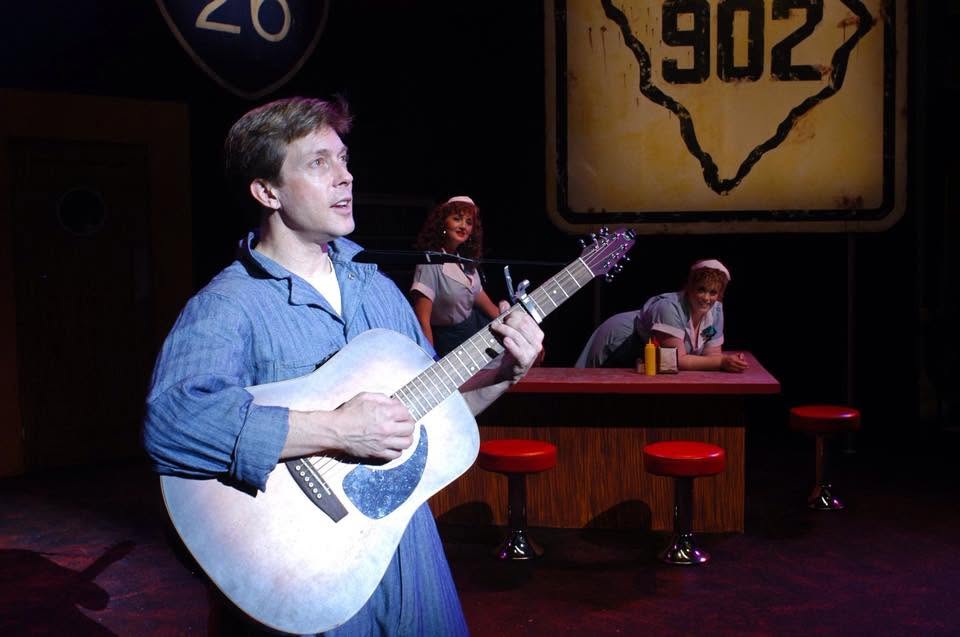Young man playing guitar on stage with a woman singing in the background, and a Texas highway sign overhead.