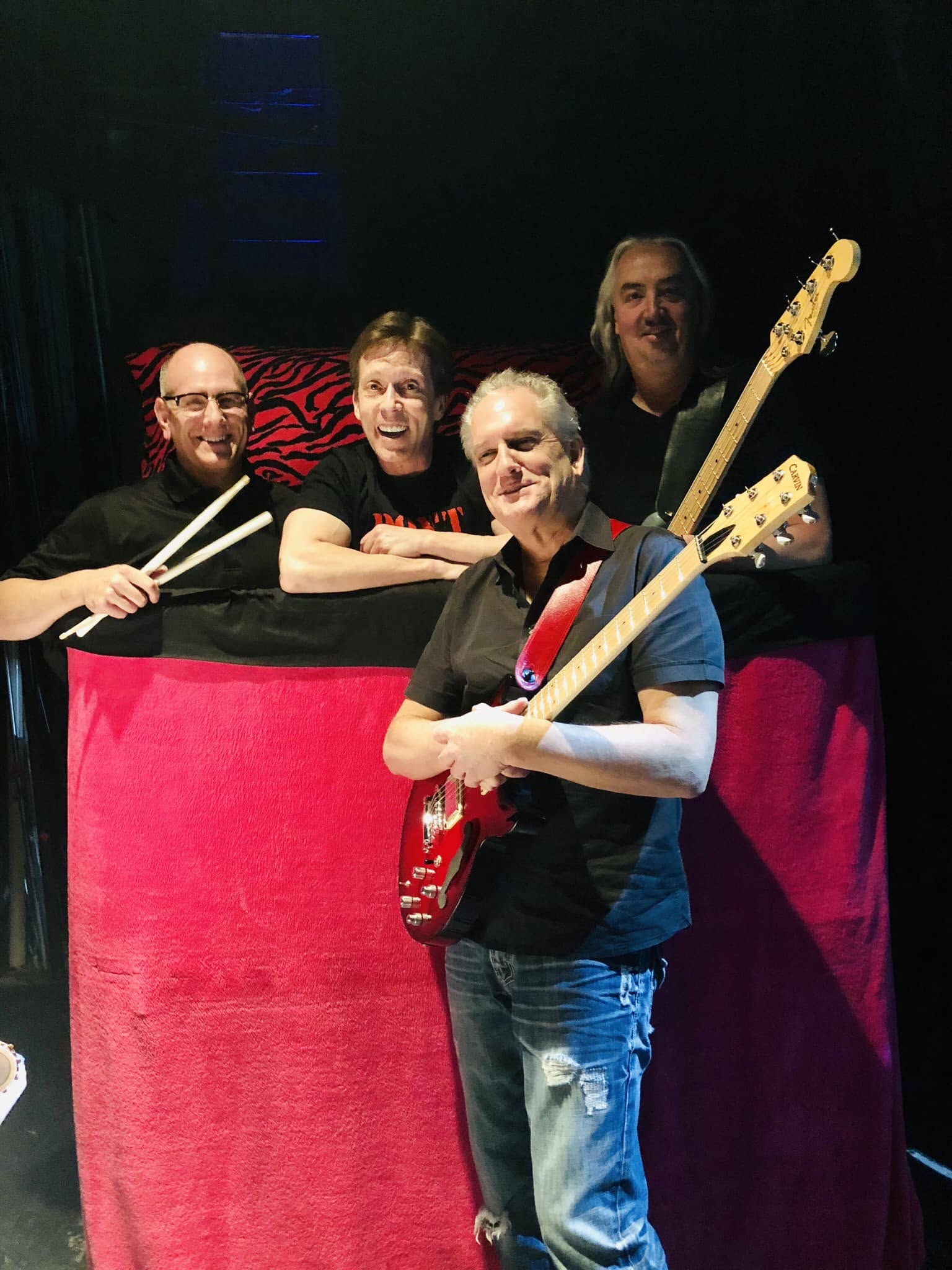 Four people on a stage, one holding a guitar, smiling, with a red curtain backdrop.
