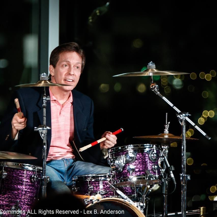 Person playing drums at night, holding drumsticks, with a cityscape background featuring blurred lights.