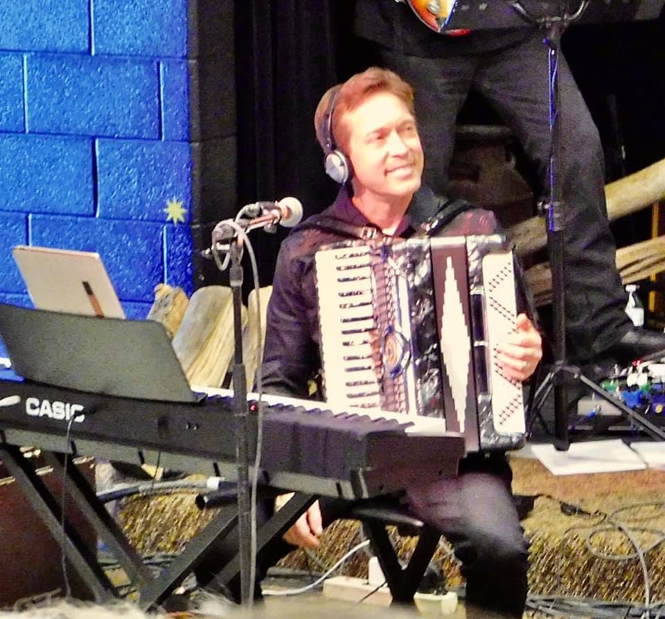 Person with headphones playing an accordion, sitting in front of a keyboard and microphone, smiling, in a performance setting.