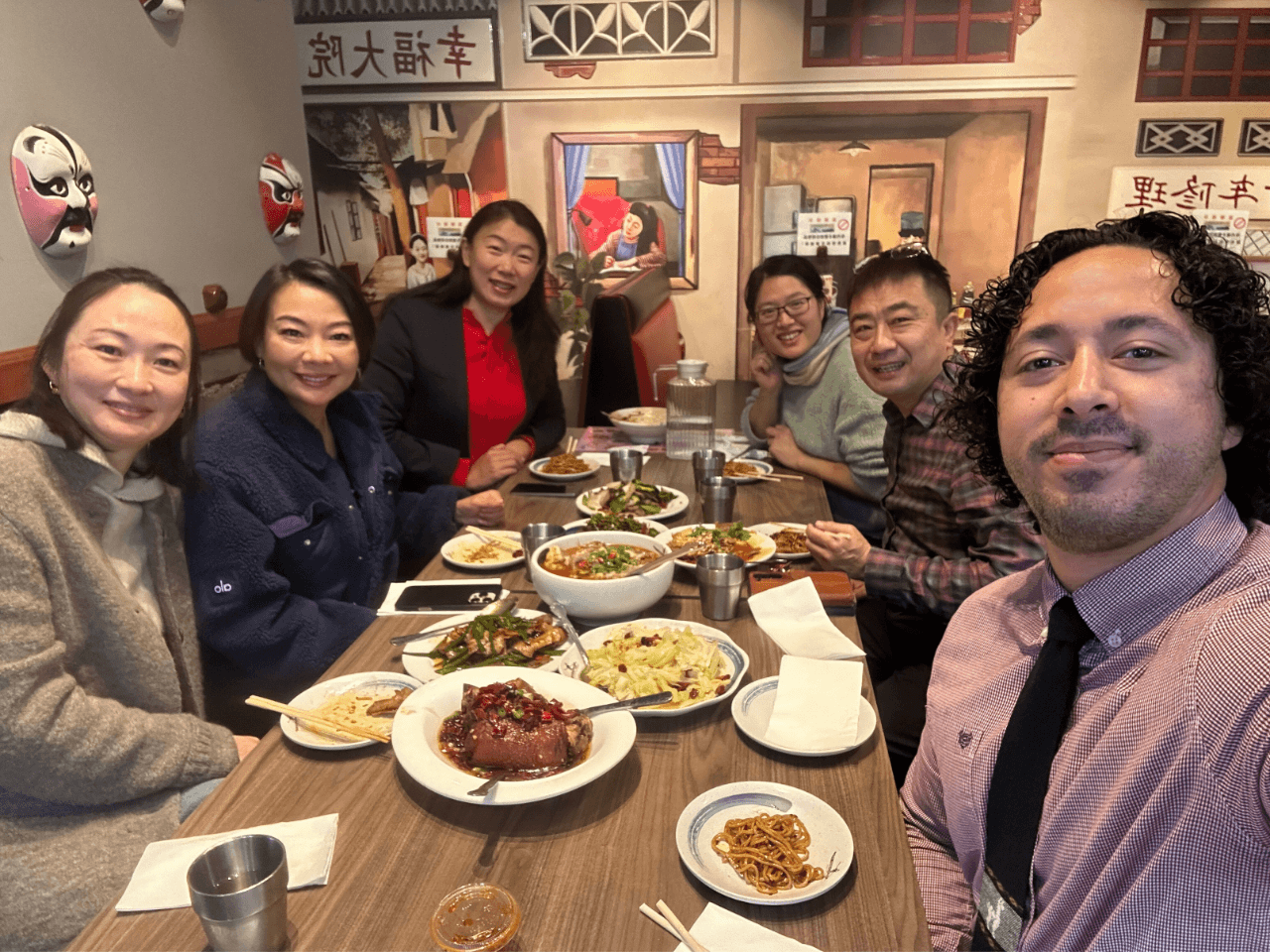 Group of six people sitting around a table with food in a restaurant, smiling at the camera.