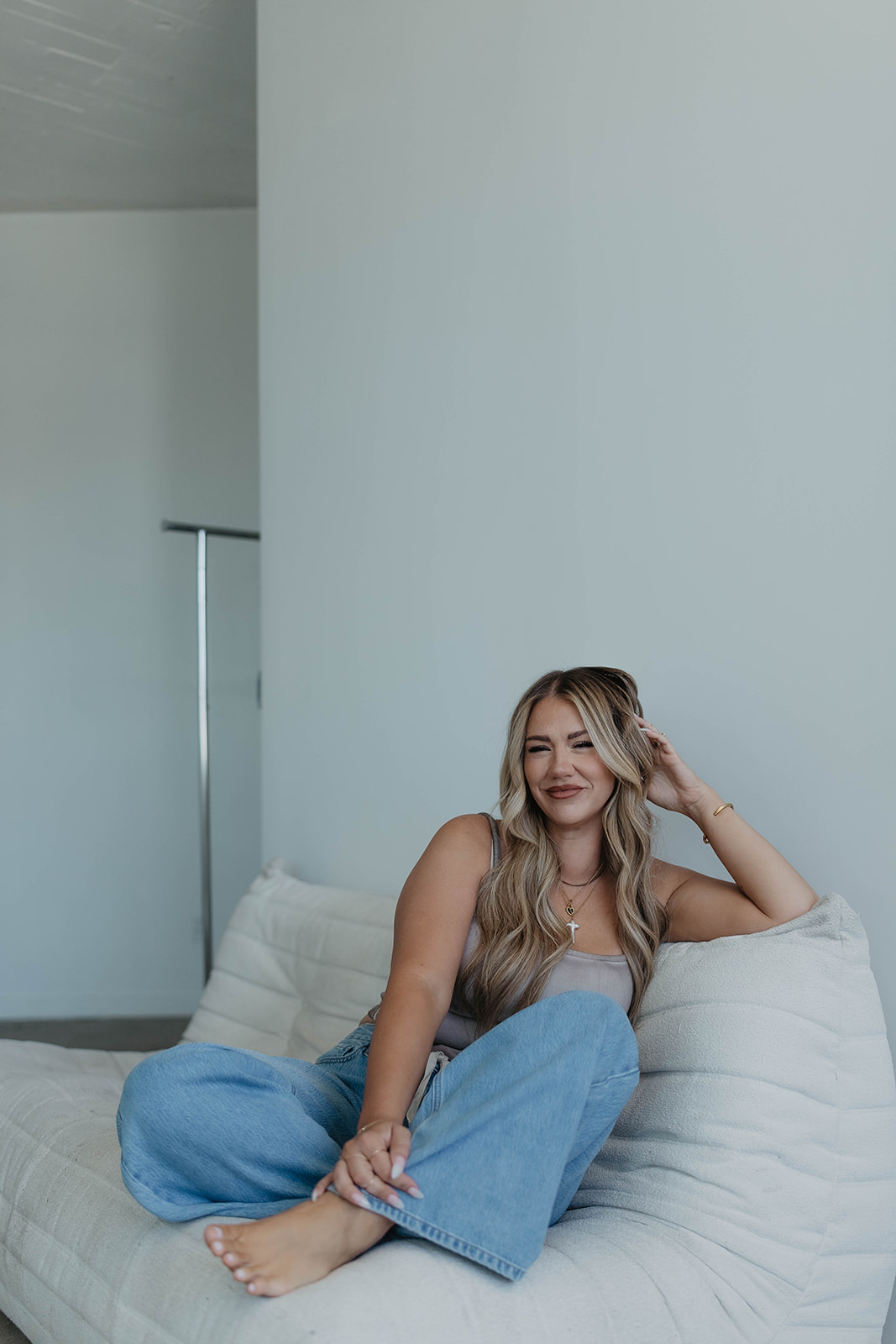Woman with long hair sitting on a bed, smiling, wearing a sleeveless top and jeans, in a minimal room.