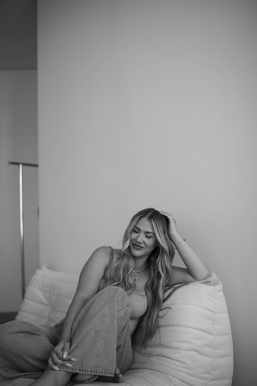 Woman with long hair sitting on a bed, resting her head on her hand, smiling, in a room with plain wall.