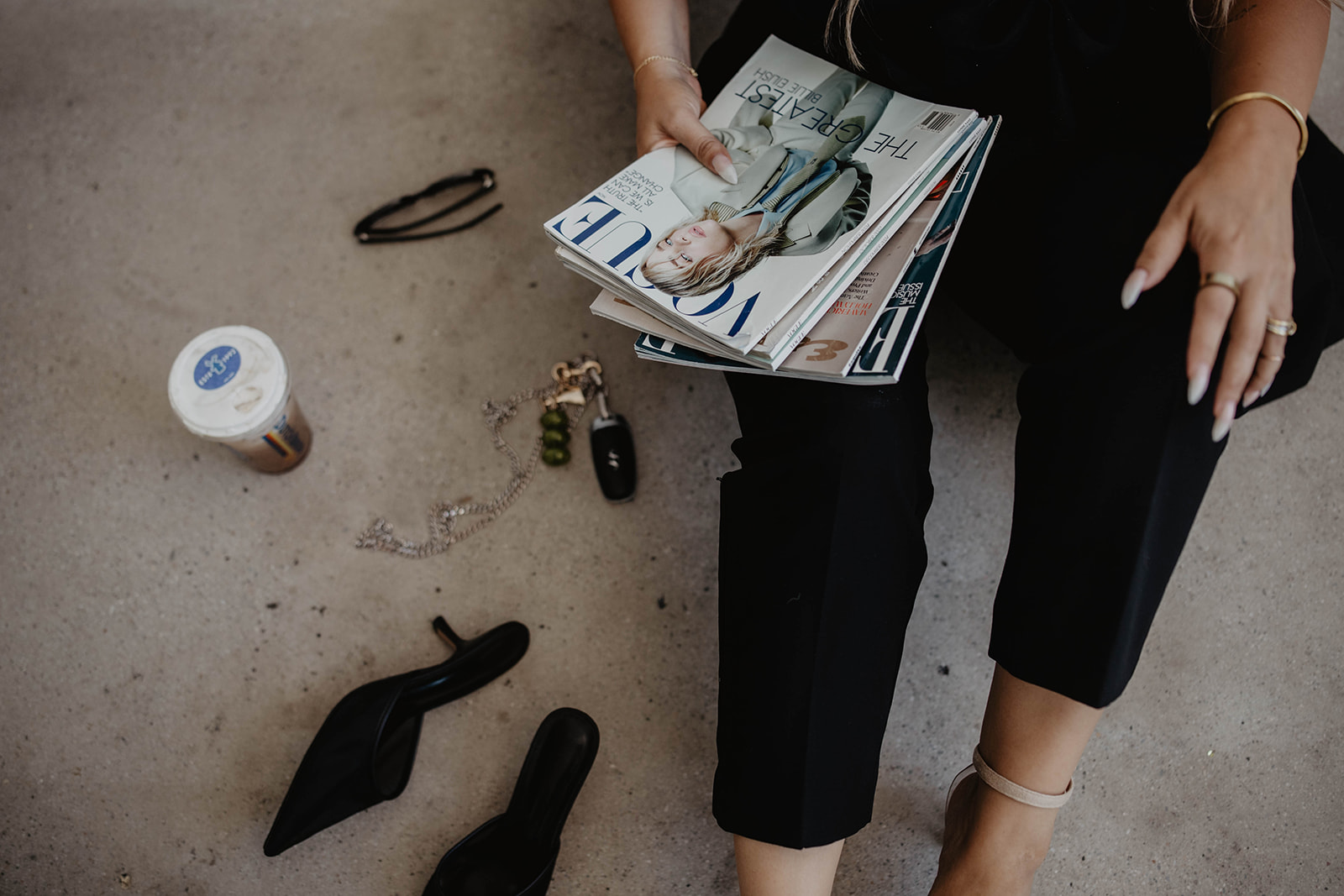 Person sitting on ground with magazines, a pair of glasses, and personal items nearby, wearing black pants and beige shoes.