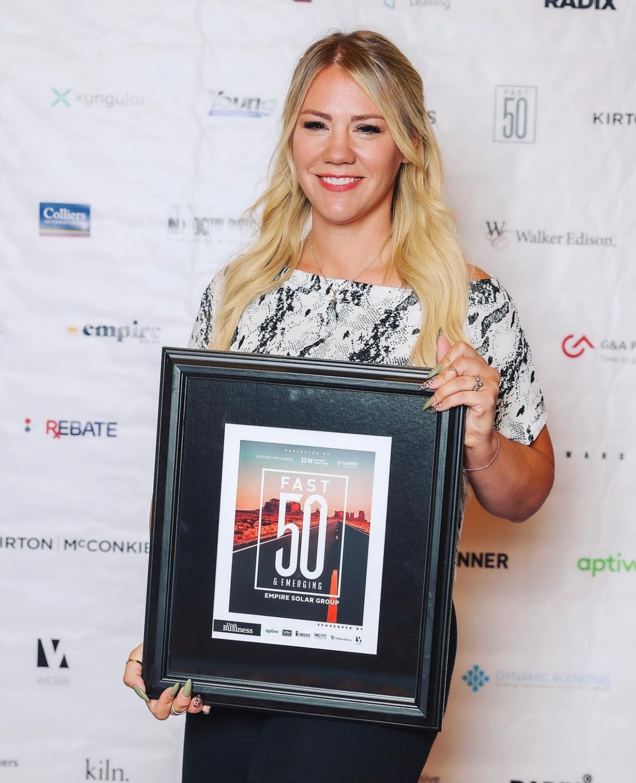 Smiling woman holding framed award with a poster inside, standing in front of a white backdrop with logos.