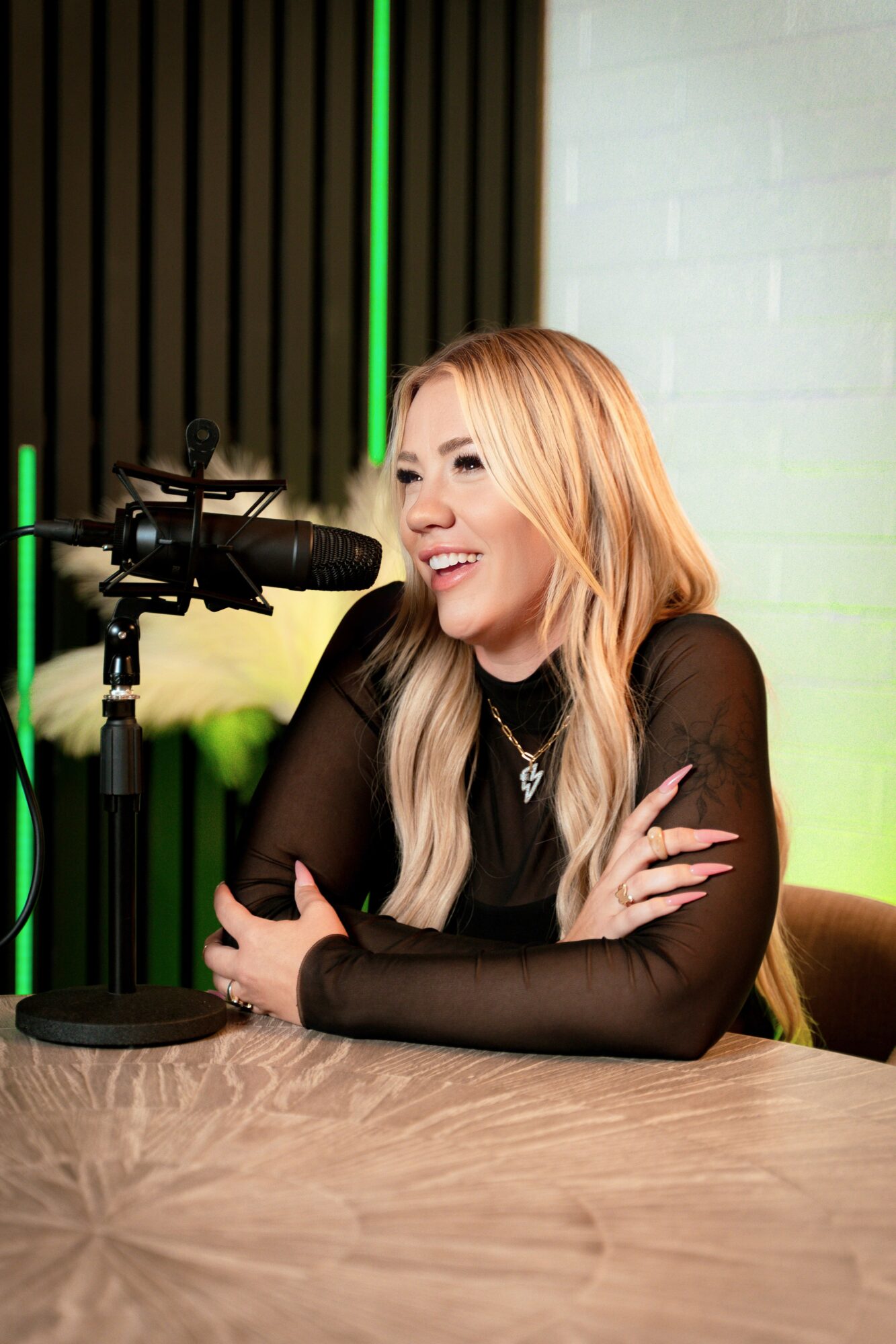 Woman with long blonde hair speaking into a microphone, smiling, sitting at a table in a studio setting.