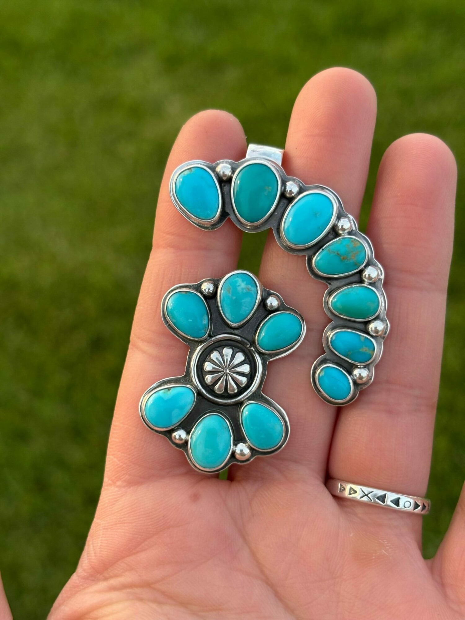 Hand holding a silver brooch with turquoise stones, a ring on finger, against a green background.