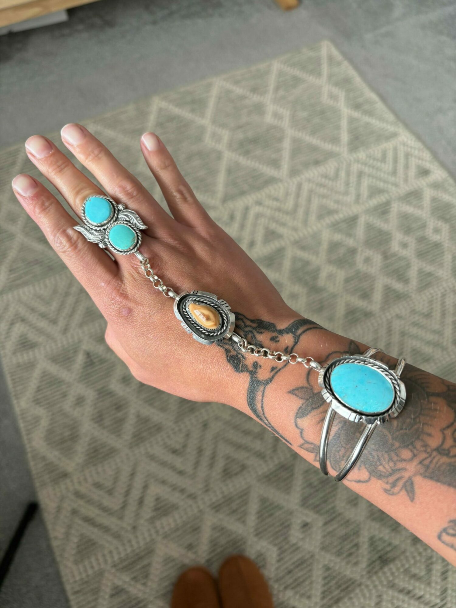 Hand with tattoos wearing a bracelet and rings, resting on a patterned surface.