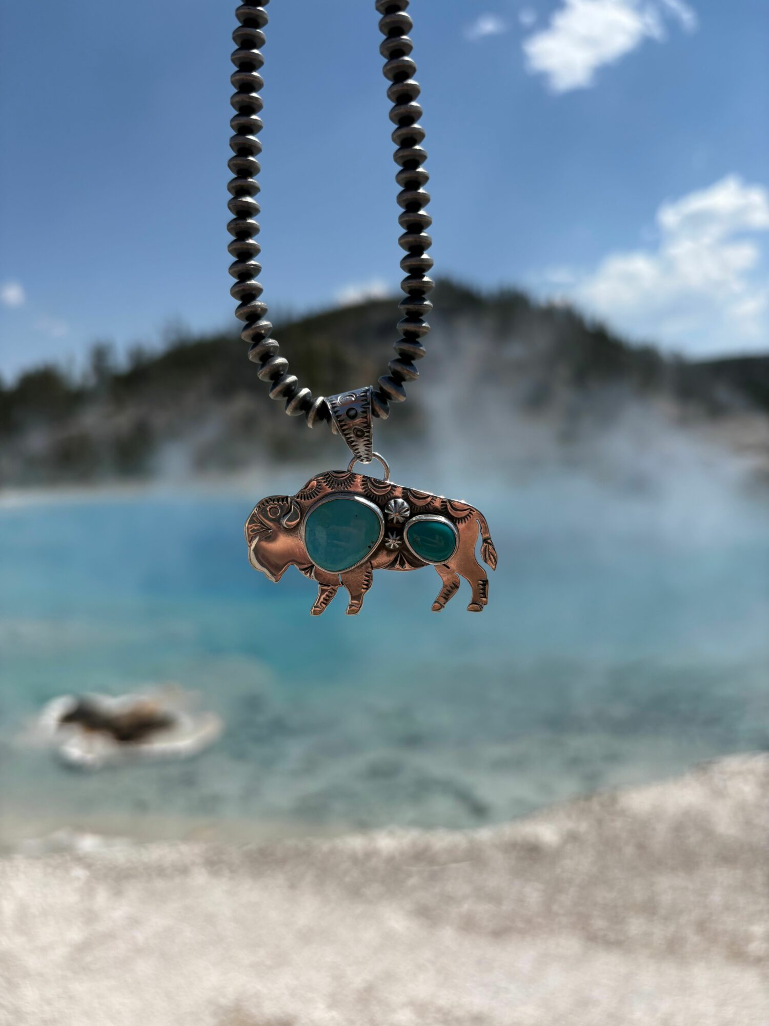 A person wearing a ring with a large blue stone, hanging from a chain, near a hot spring with steam and rocks.