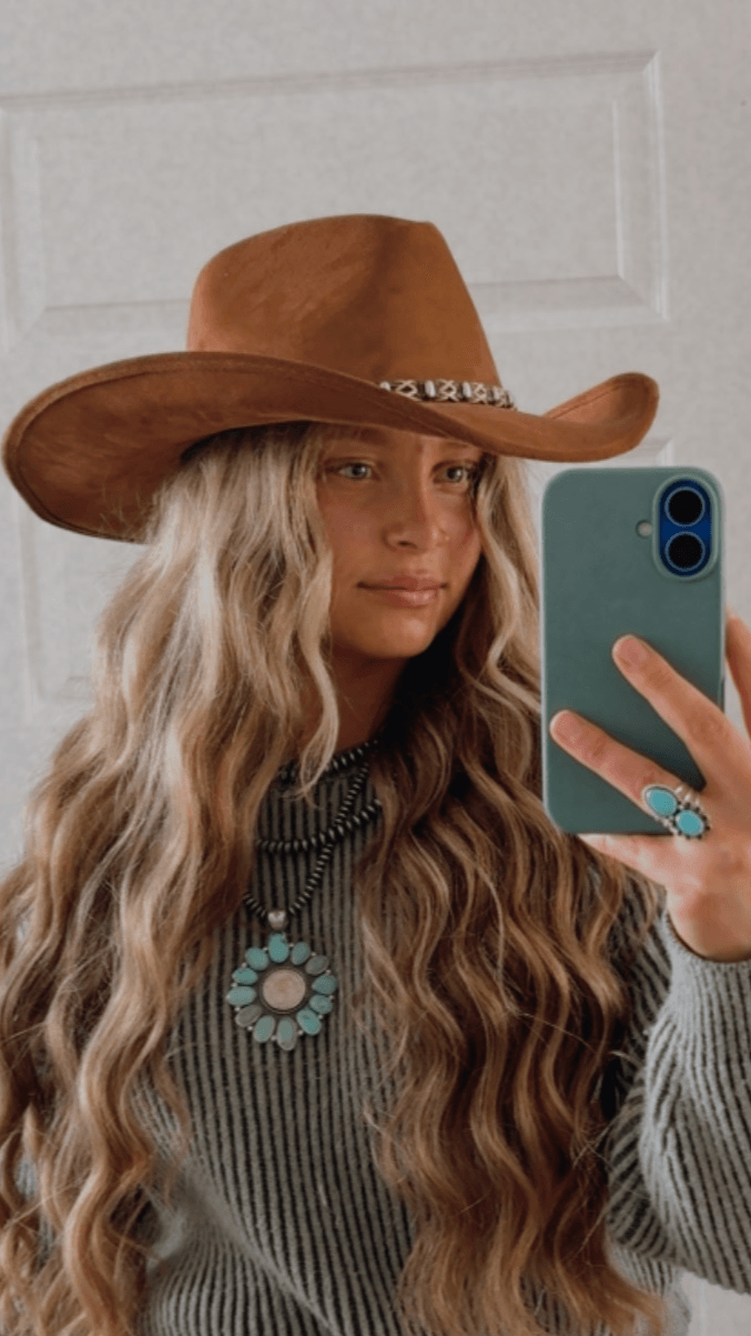 Woman with long wavy hair wearing a cowboy hat and striped sweater, taking a selfie with a smartphone.