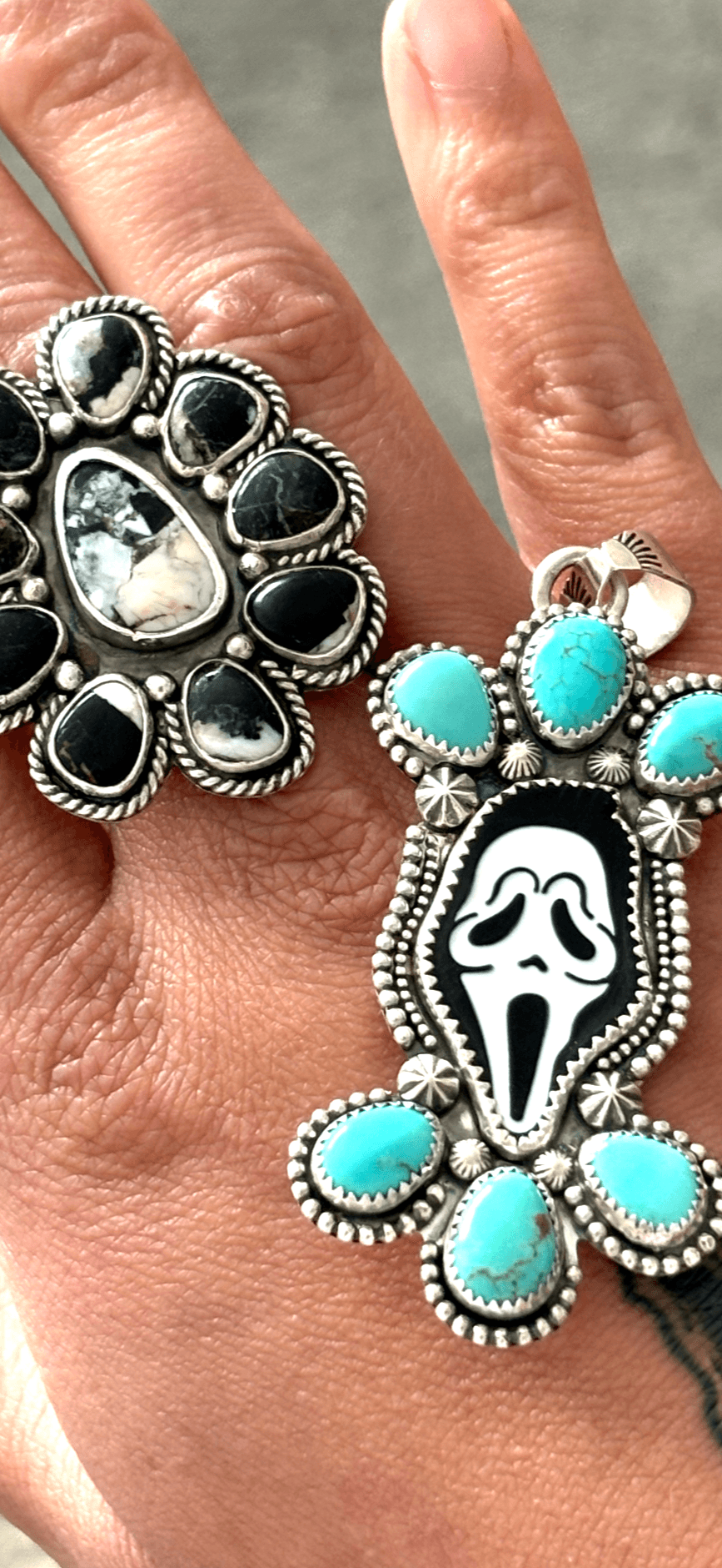 Two large rings with black, turquoise, and silver designs worn on fingers, one with a skull motif, on a hand.