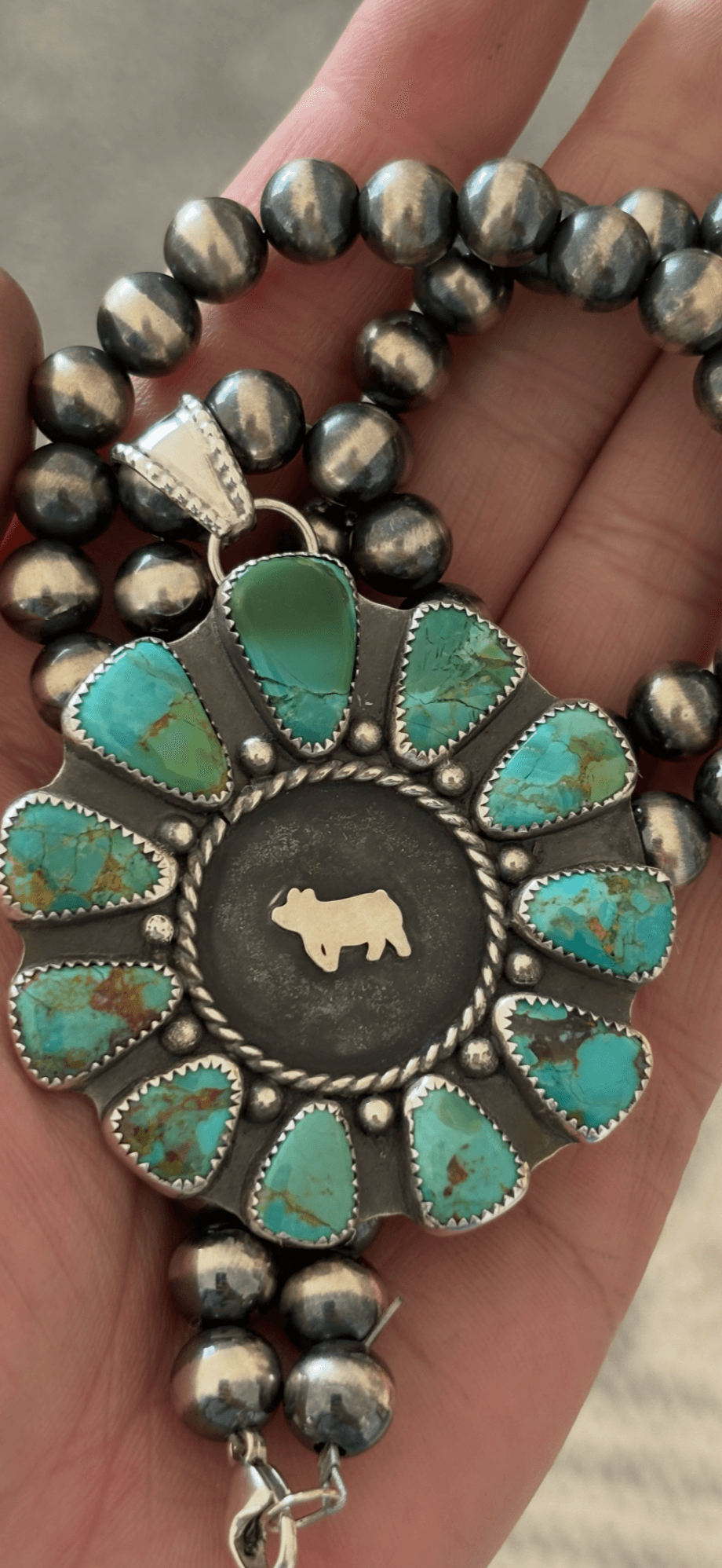 Hand holding a beaded necklace with a circular pendant featuring turquoise stones and a central animal figure, against a neutral background.