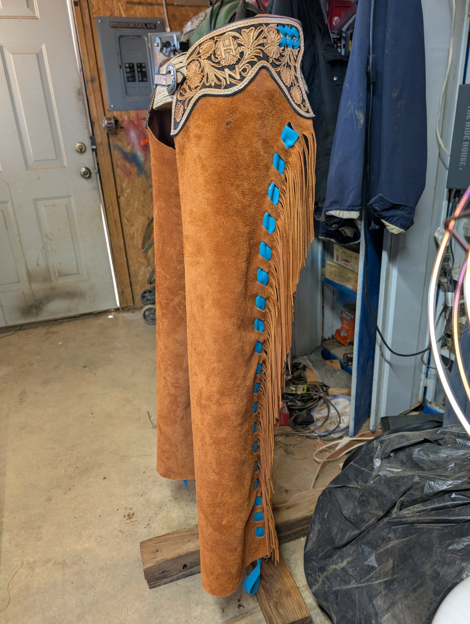 Cowboy chaps with decorative embroidery and blue accents hanging in a garage, with a door and various tools visible in the background.