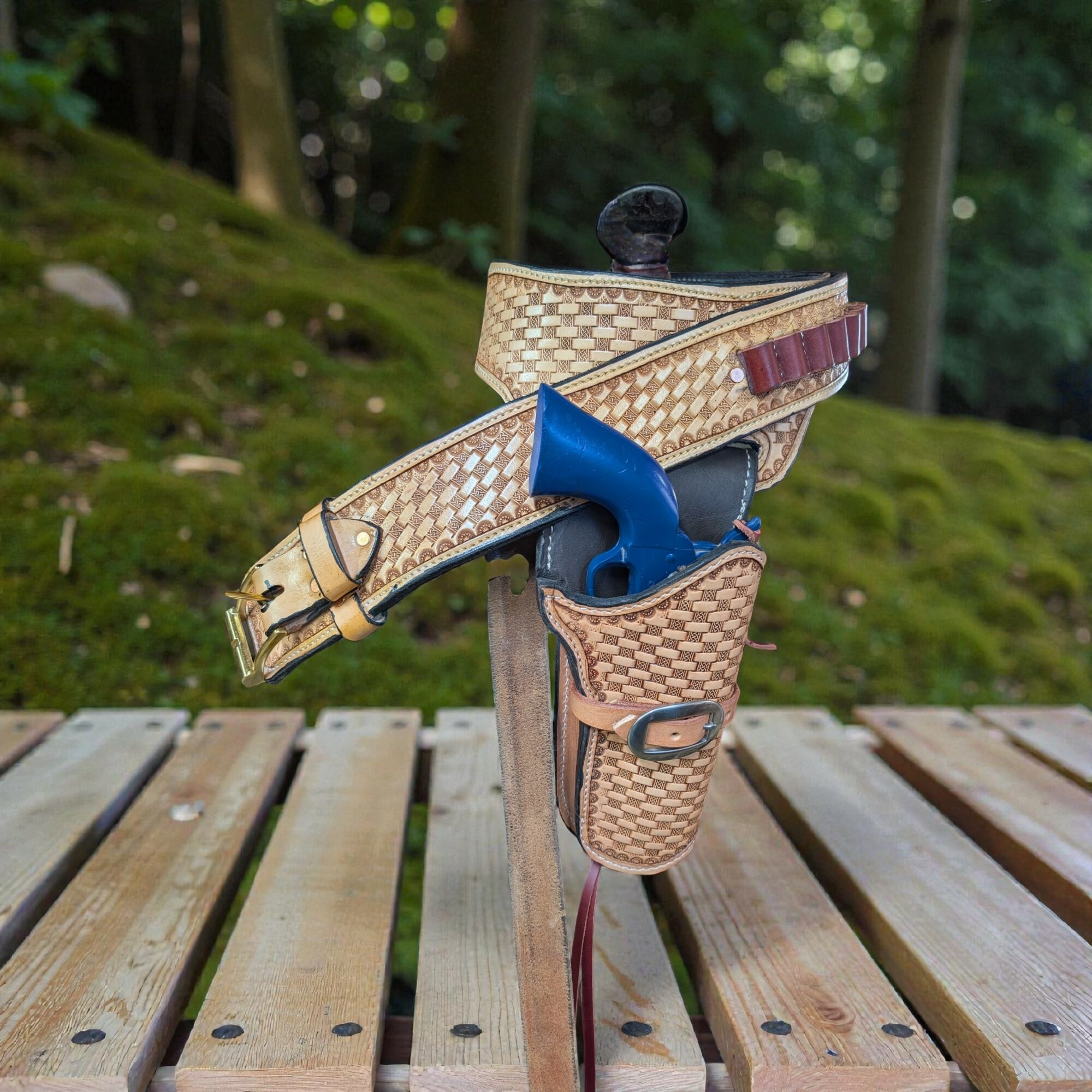 A handgun with a beige patterned holster and blue grip, placed on a wooden surface outdoors with trees in the background.