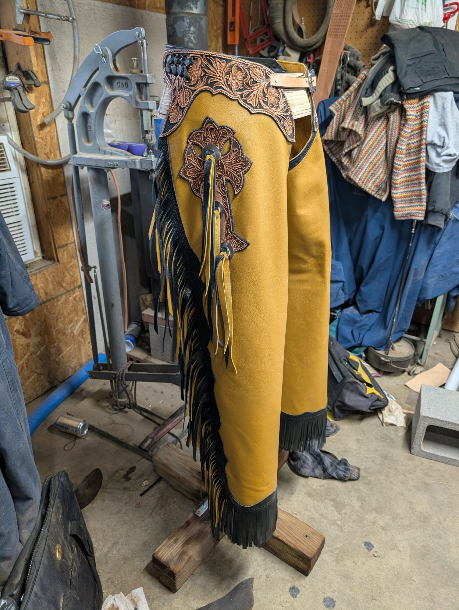 A pair of yellow chaps with black fringe and decorative embroidery, hanging in a cluttered workshop.