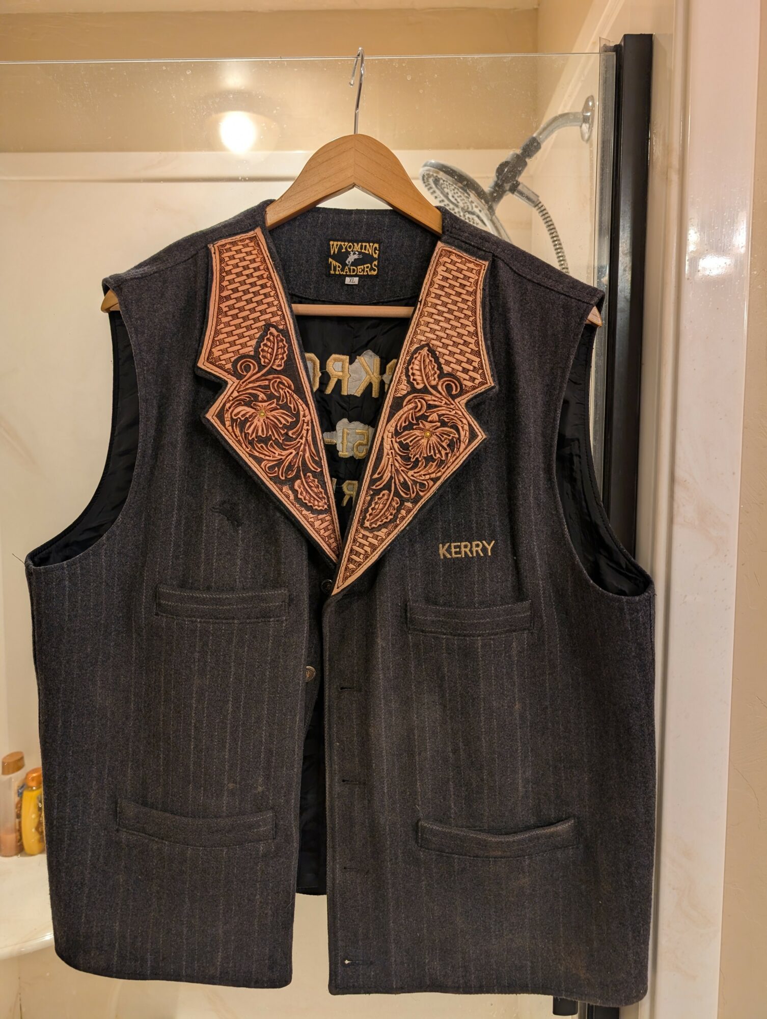 Dark vest with embroidered collar hanging on a hanger, with a showerhead visible in the background.