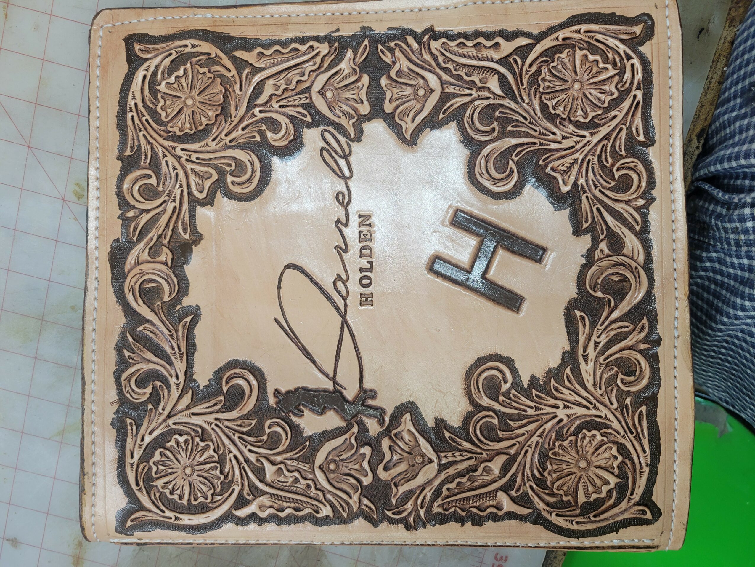 Decorative wooden plaque with floral patterns, signature, and the letter H, viewed sideways.