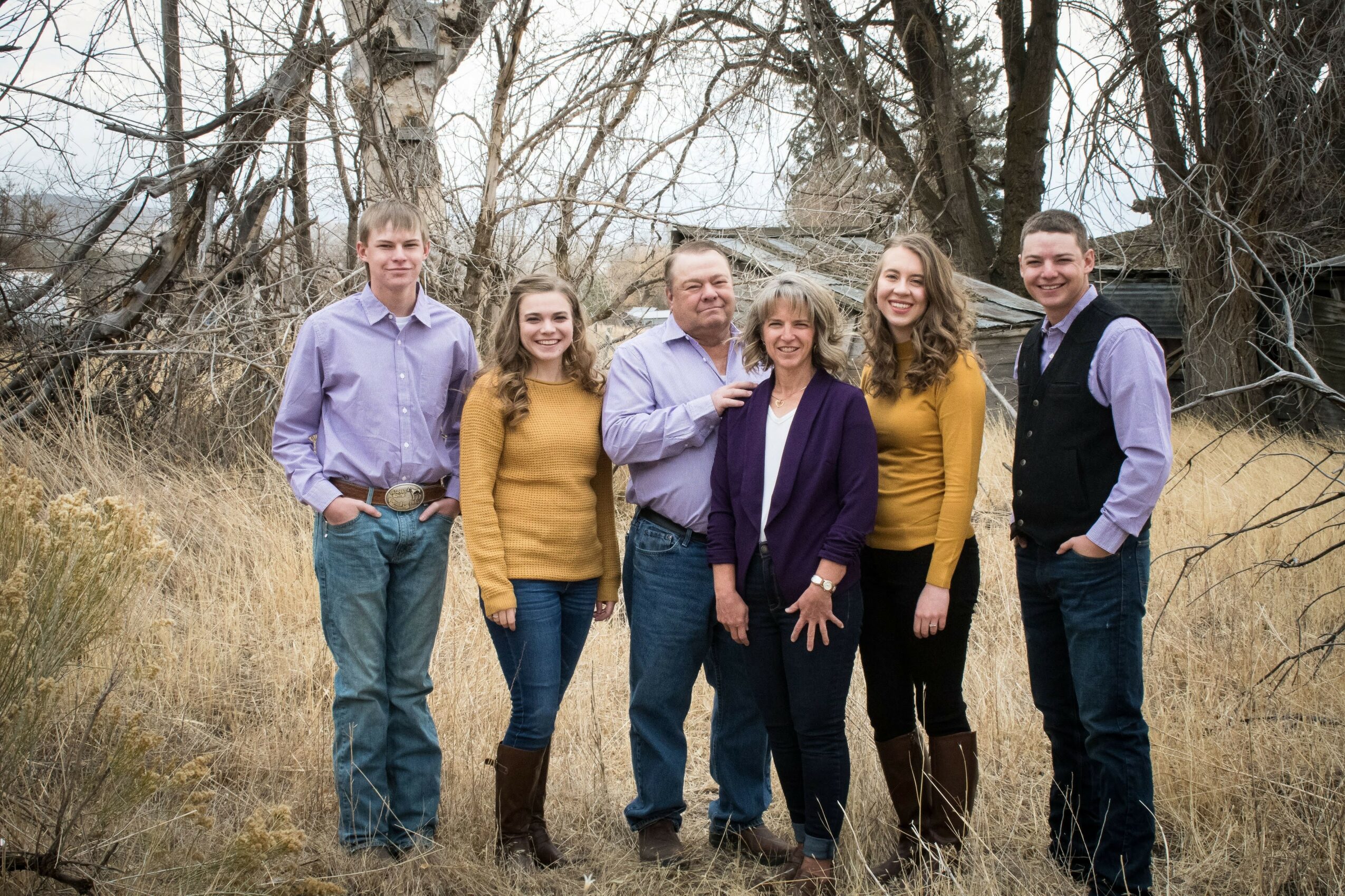 Group of six people standing outdoors in a grassy area with trees, smiling, dressed in casual clothing, autumn setting.