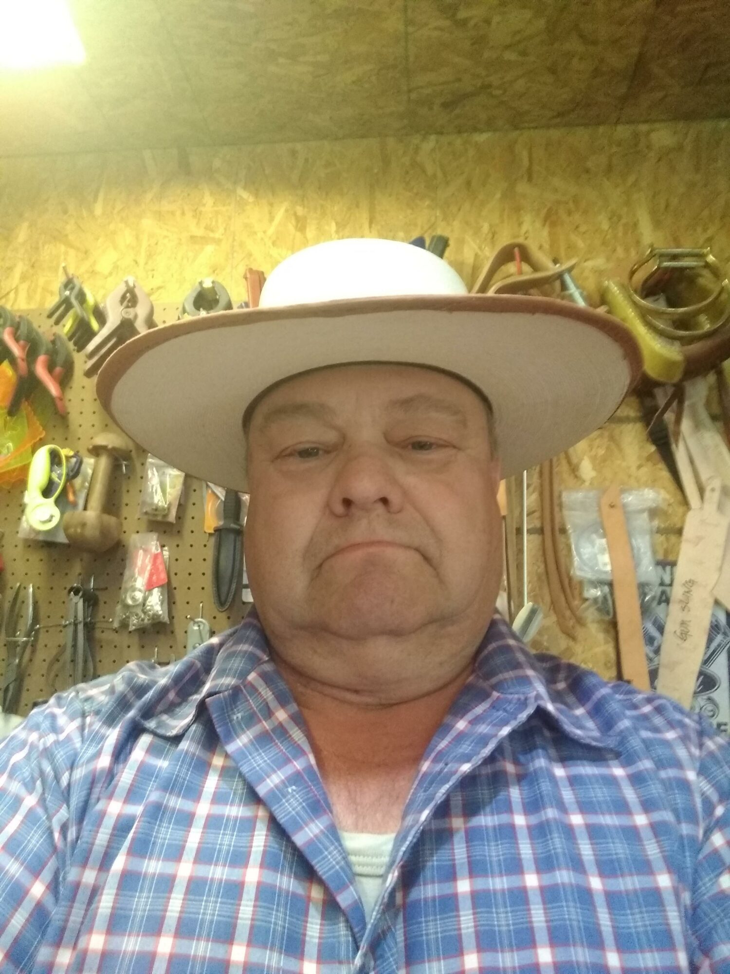 Man wearing a wide-brimmed hat and plaid shirt, standing in a workshop with tools on the wall behind him.