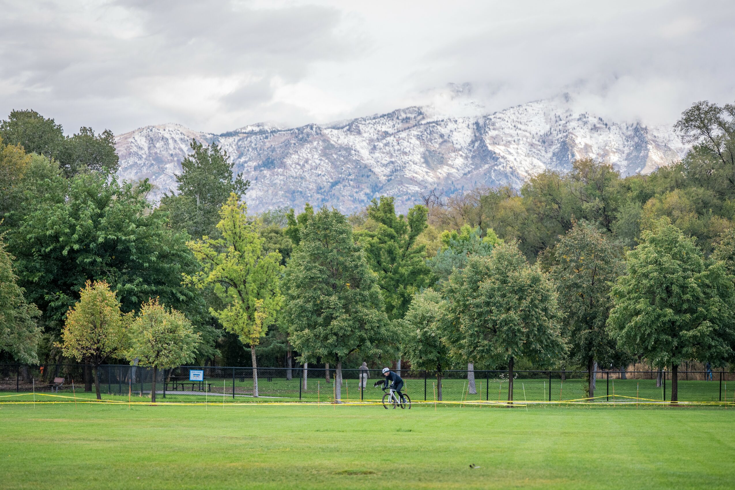 Park with trees, a person riding a bicycle, and mountains in the background, under cloudy sky.