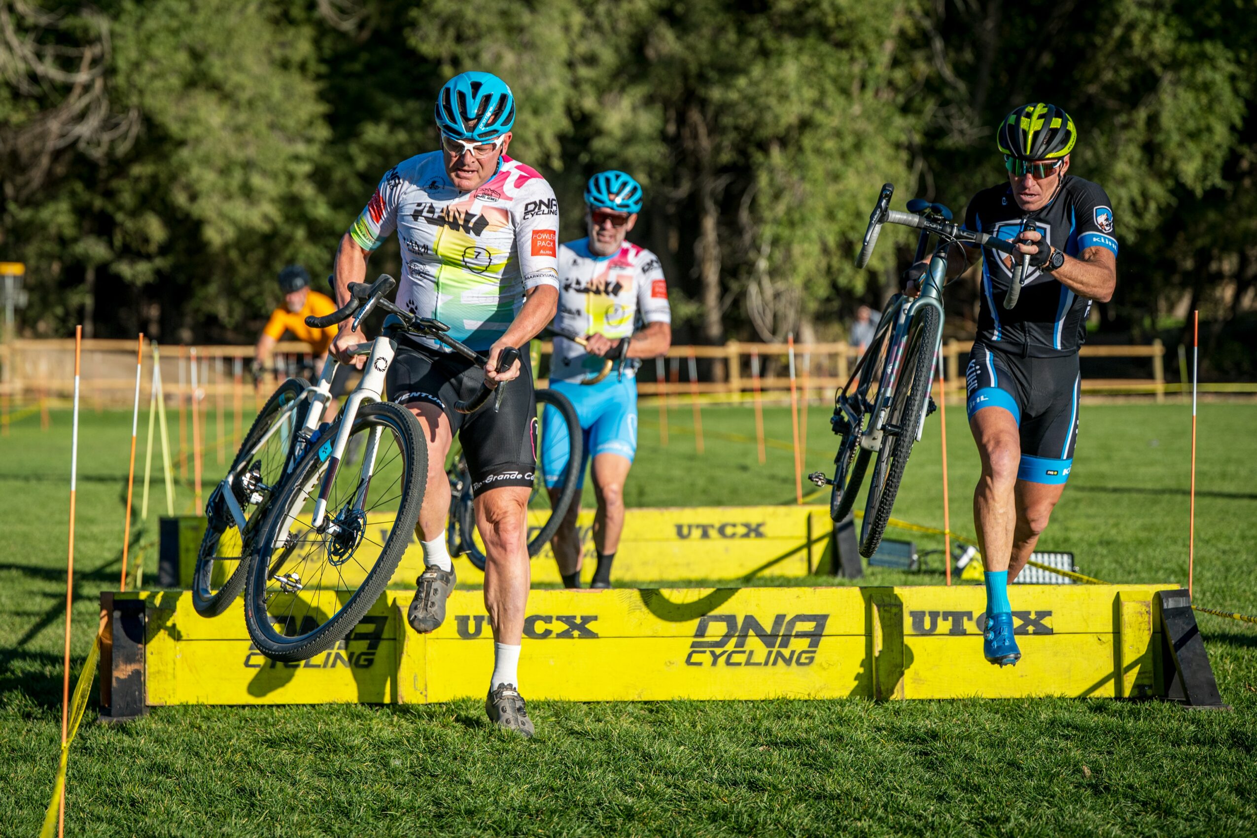 Three cyclists crossing a yellow barrier during a race, with trees in the background, one lifting a bike over the barrier.