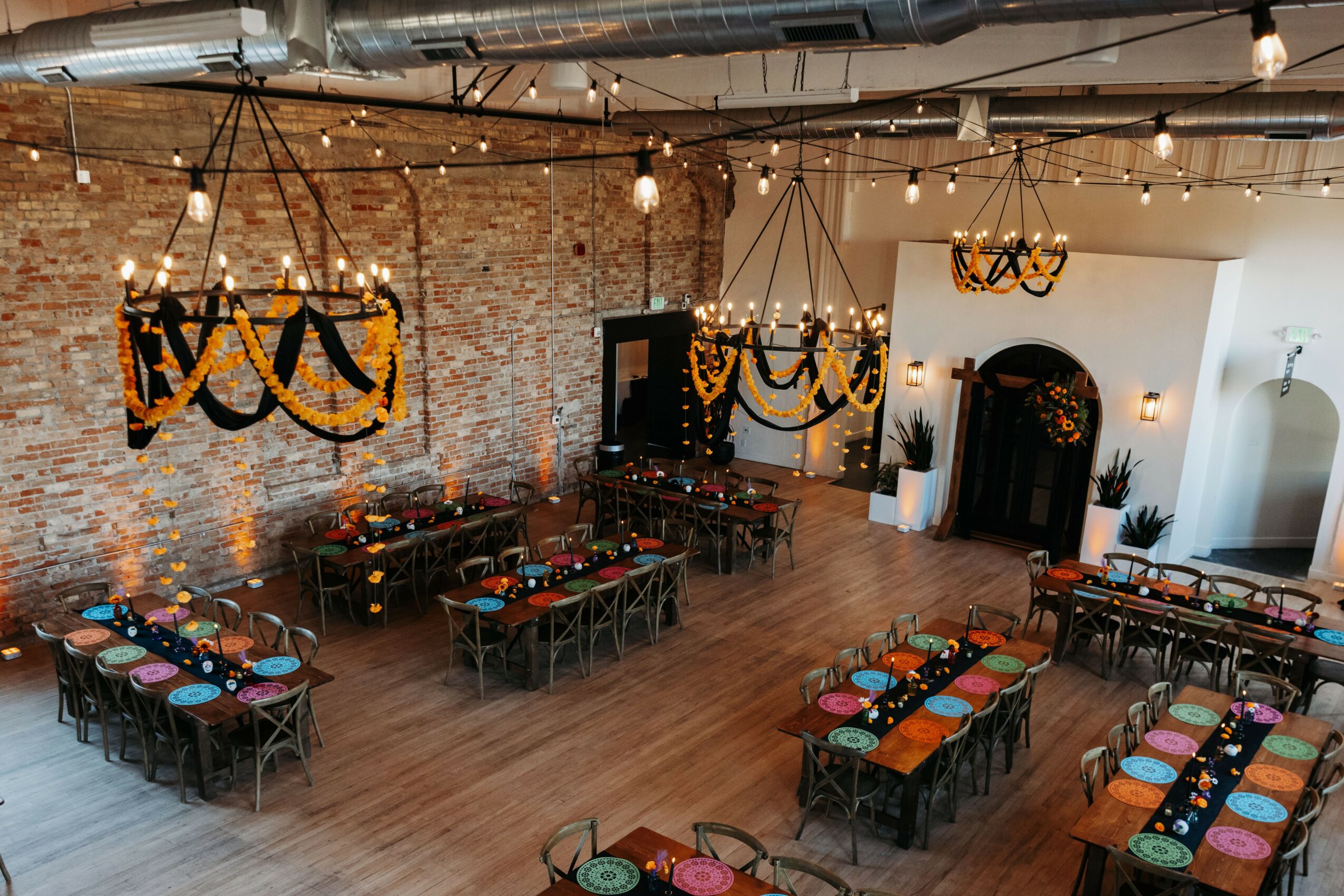 Event space with brick wall, string lights, and decorated tables with colorful plates and napkins, wooden floor, and arched doorway.