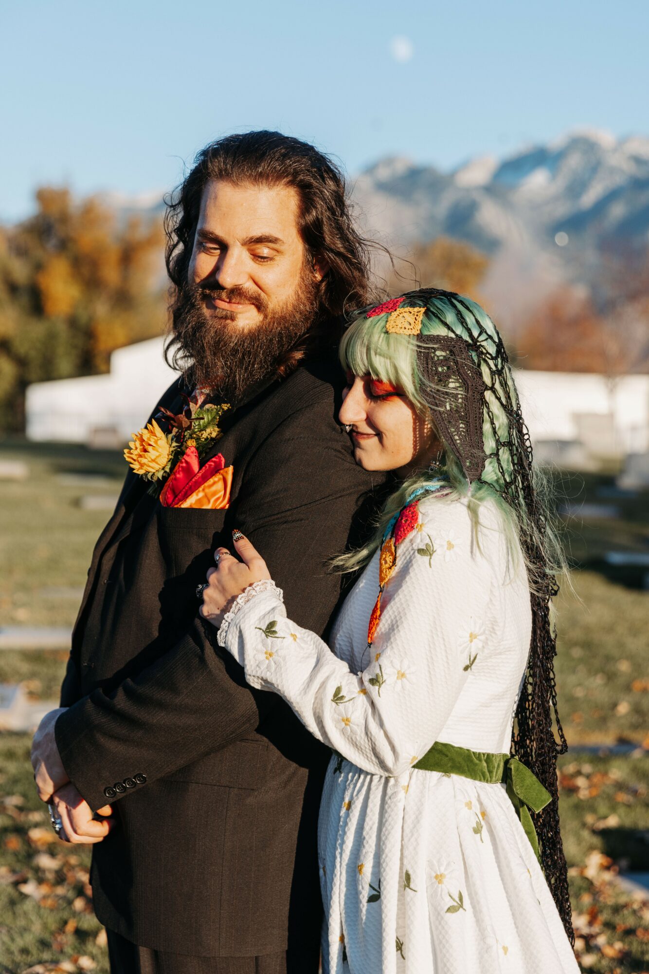 A man with long hair and a beard in a black suit and a woman with green hair and a white dress stand outdoors with mountains in the background.